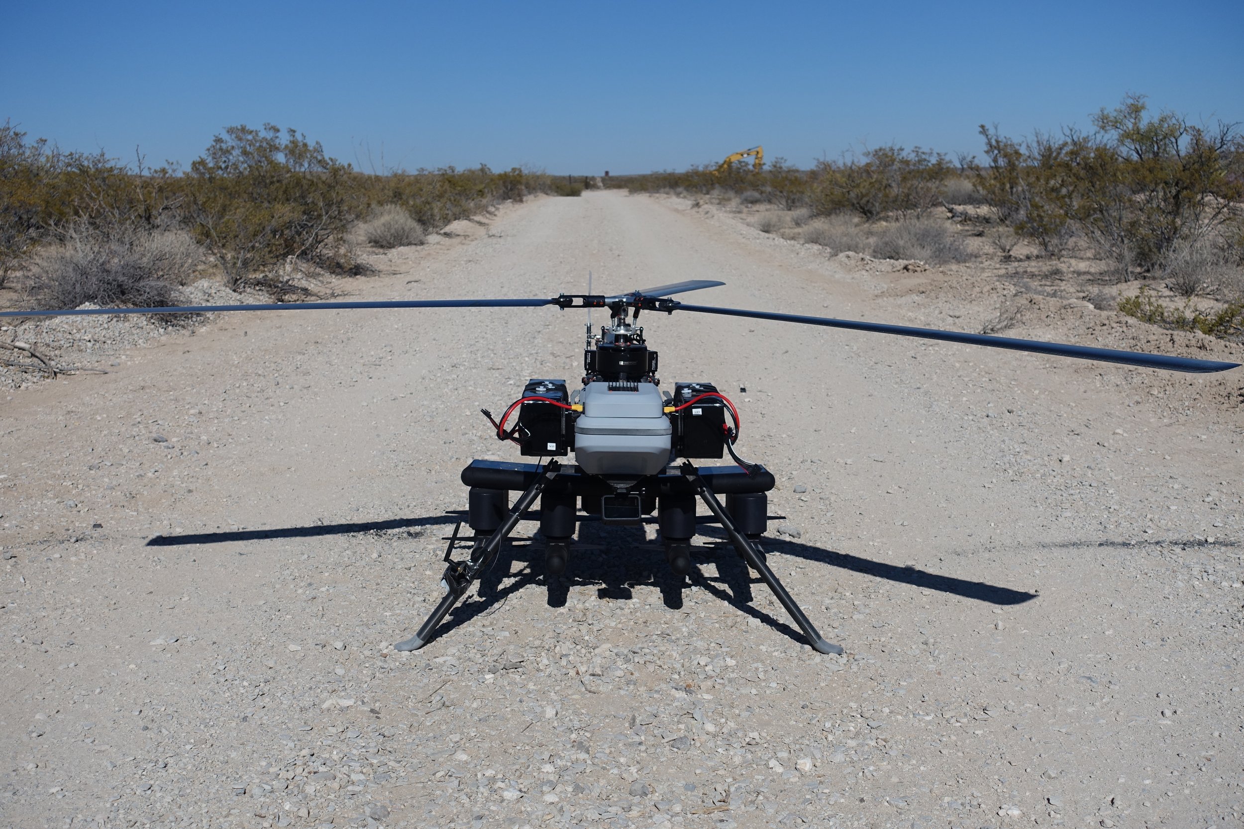 A drone with a large rotor on a dirt road in a desert landscape with sparse bushes, under a clear blue sky.