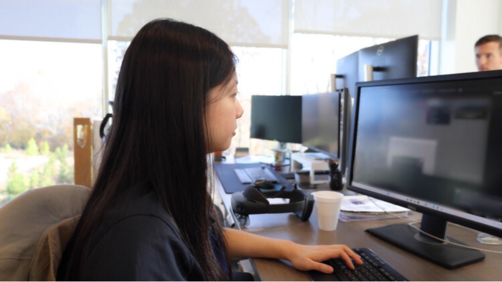 Woman working at a desk with multiple monitors in an office.