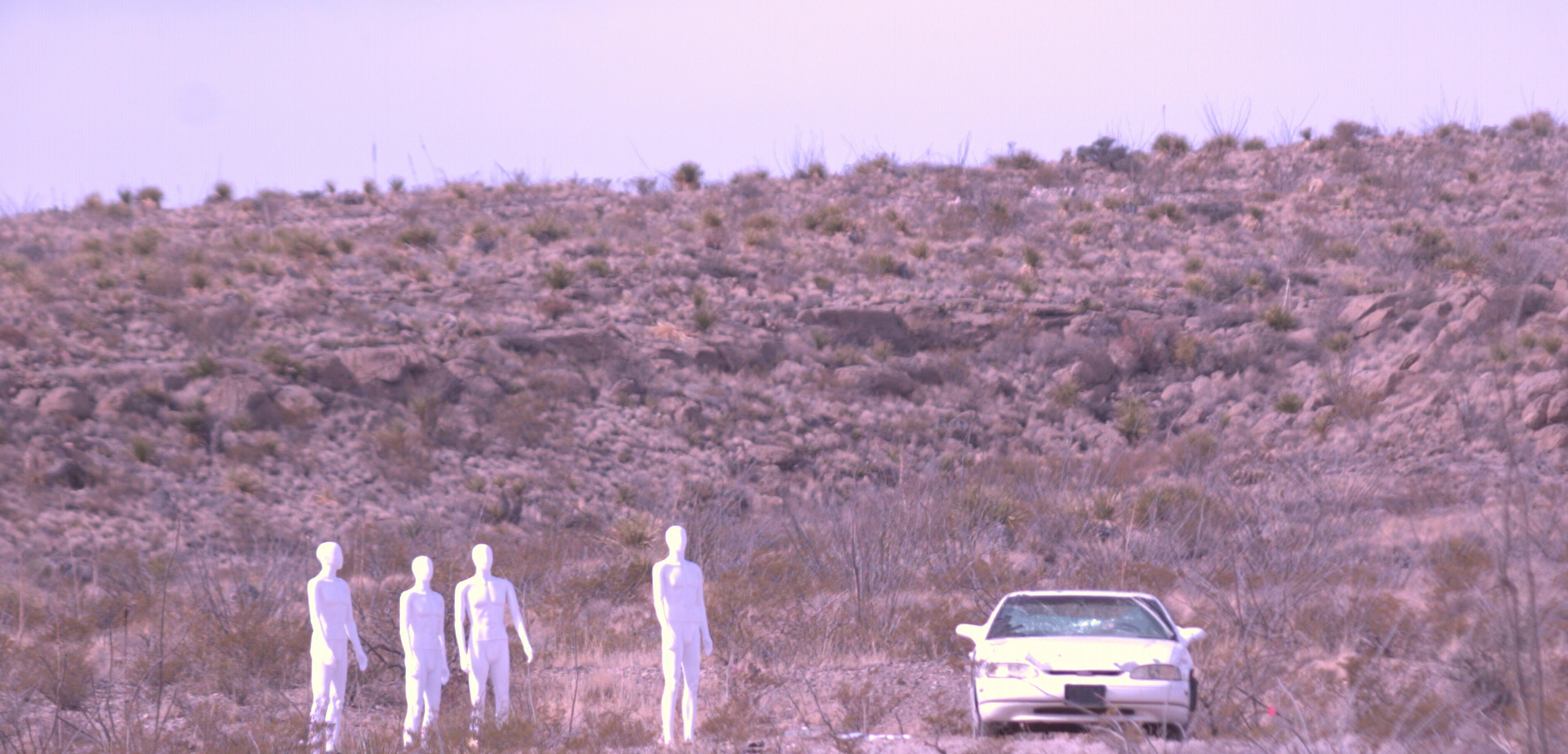 Four white mannequins standing in a barren desert landscape with a white car nearby, set against a backdrop of rocky hills and sparse desert vegetation.