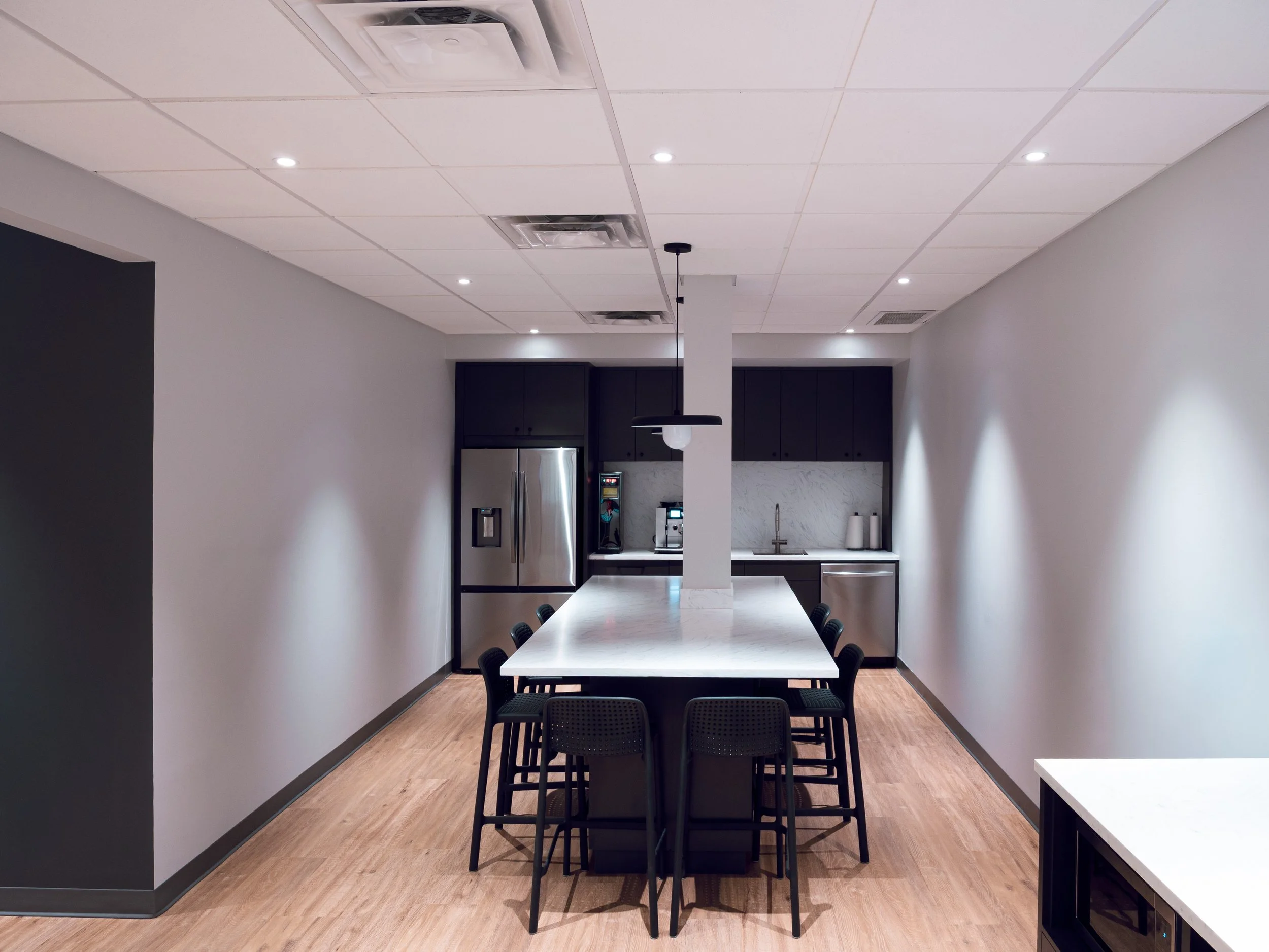 Modern kitchen with a large central island, black chairs, and stainless steel appliances on light wood flooring.
