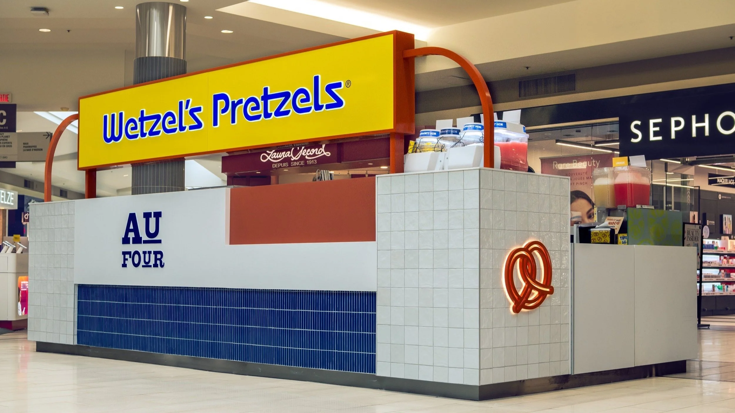 A pretzel kiosk named Wetzel's Pretzels located inside a shopping mall, with a bright yellow sign and colorful containers of pink beverage on display.
