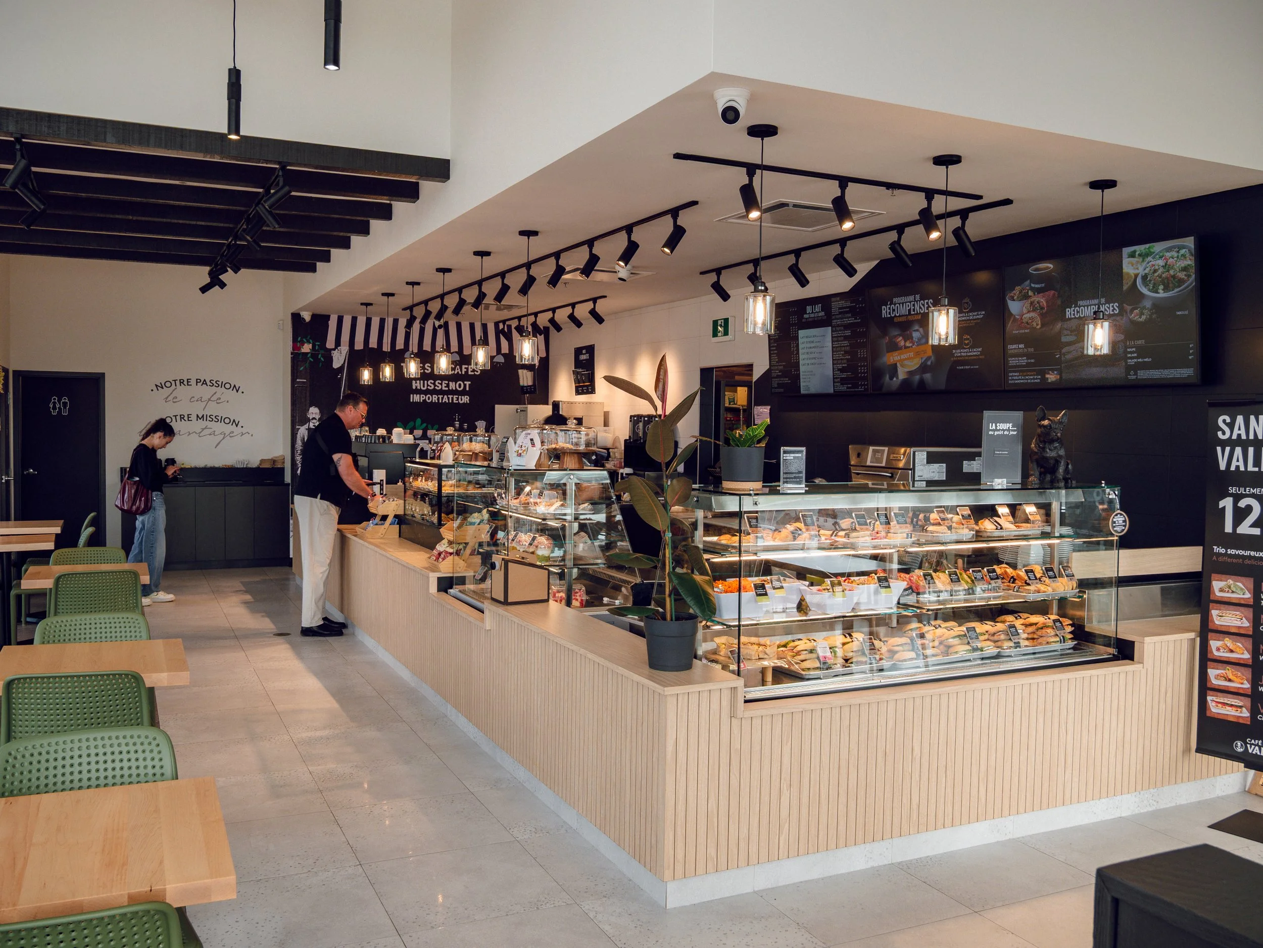 Interior of a modern cafe with a wooden display case filled with food, a menu board above, and a few customers ordering at the counter.