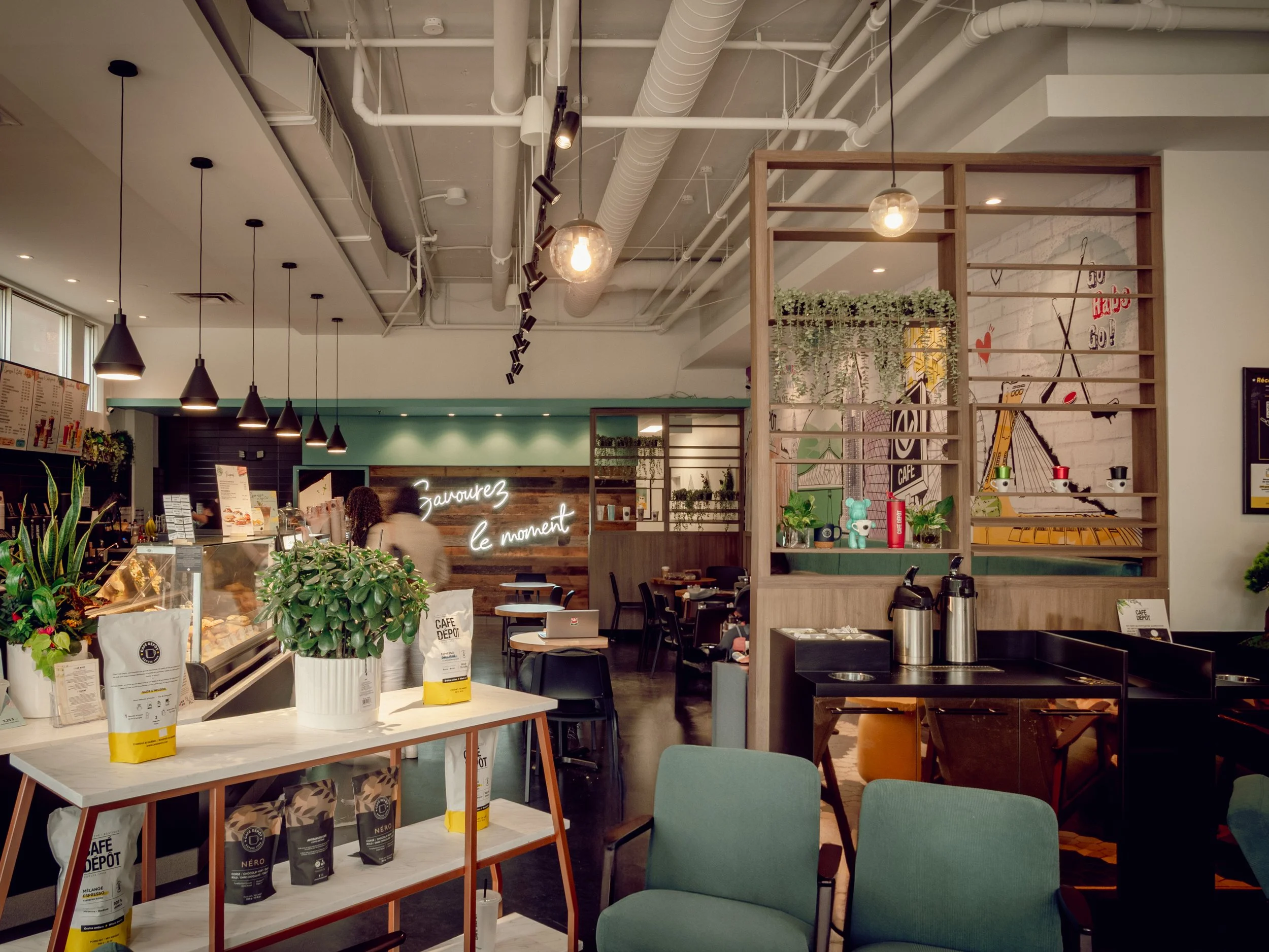 Interior of a modern coffee shop with seating, decorative plants, and a neon sign that reads 'Zaoirez le moment' in the background.