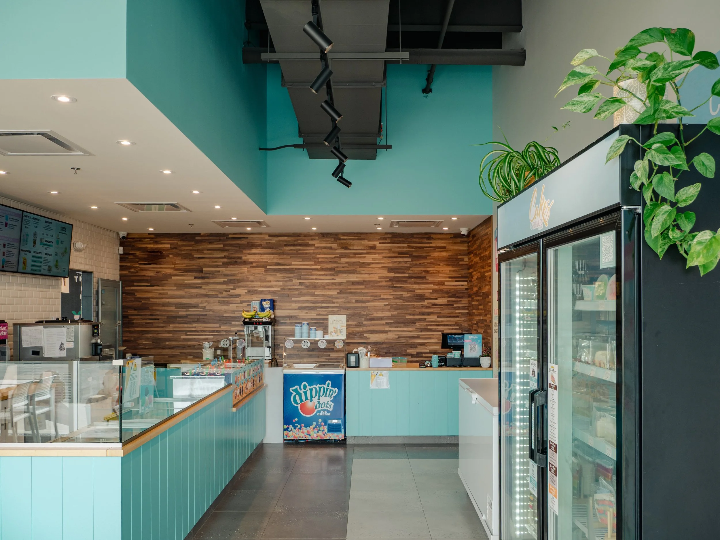 Interior of a modern ice cream shop with teal accents, a wooden accent wall, refrigerated display case, digital menus, and a small ice cream freezer.
