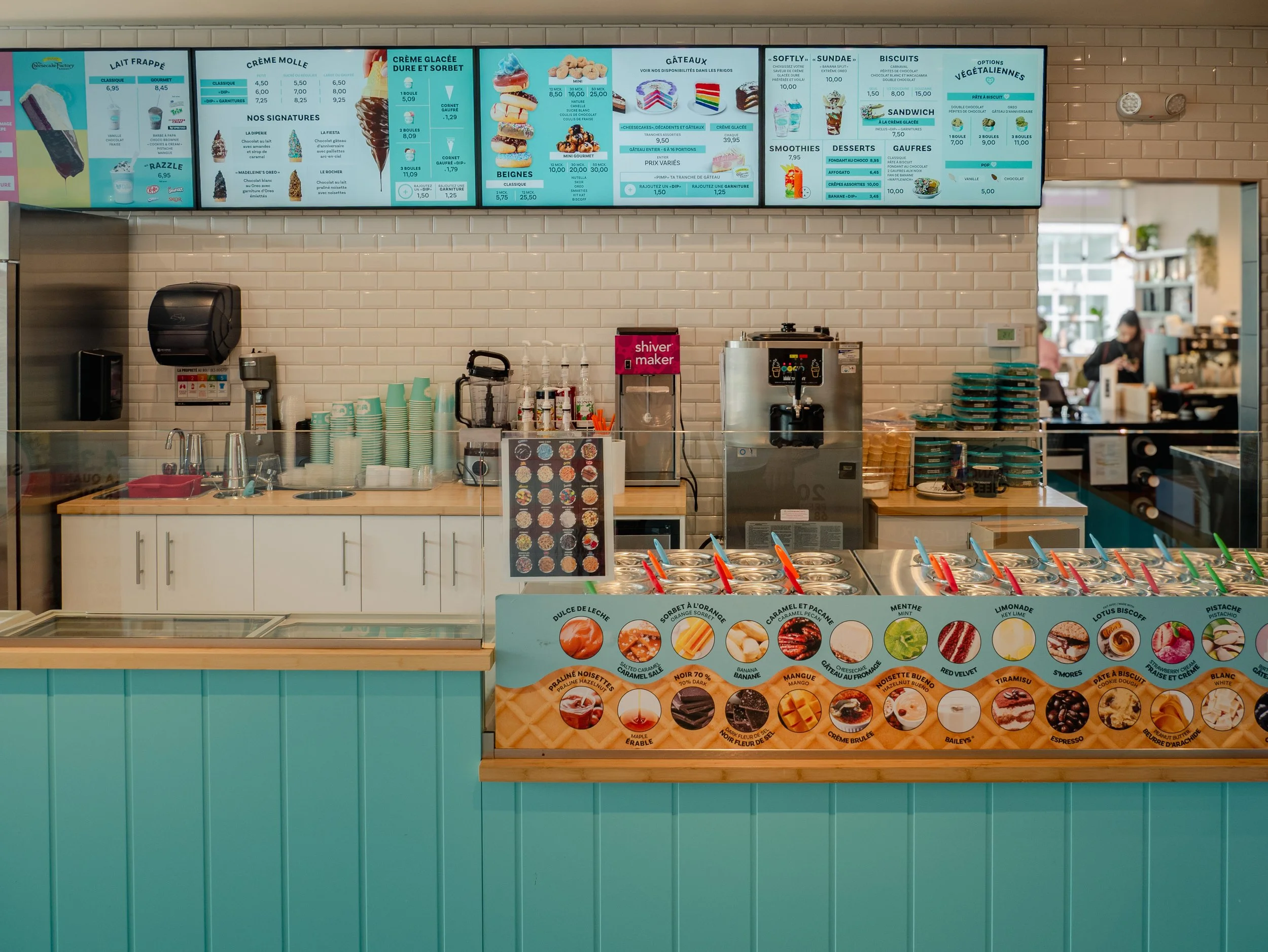 Ice cream shop counter with various flavors and toppings, digital menu above with desserts and beverages, cups, ice cream machines, and staff in the background.