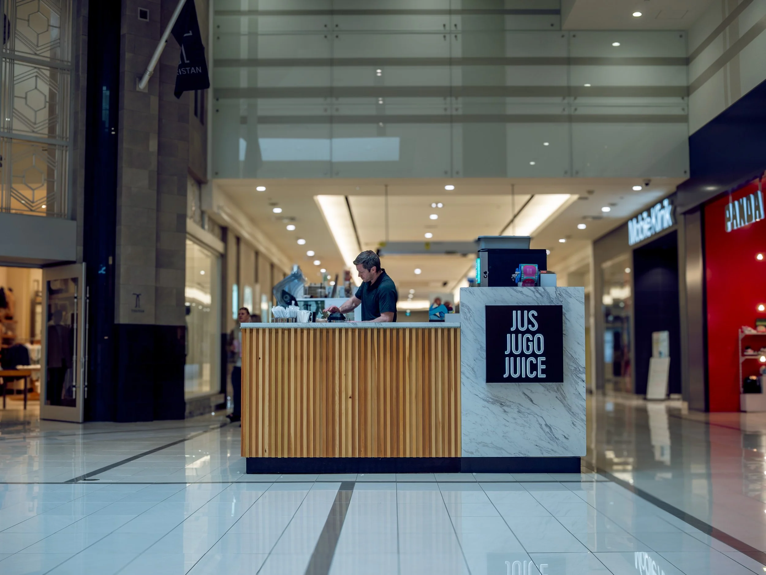 A juice stand in a shopping mall with a man working behind the counter and a sign that says 'JUS JUGO JUICE'.