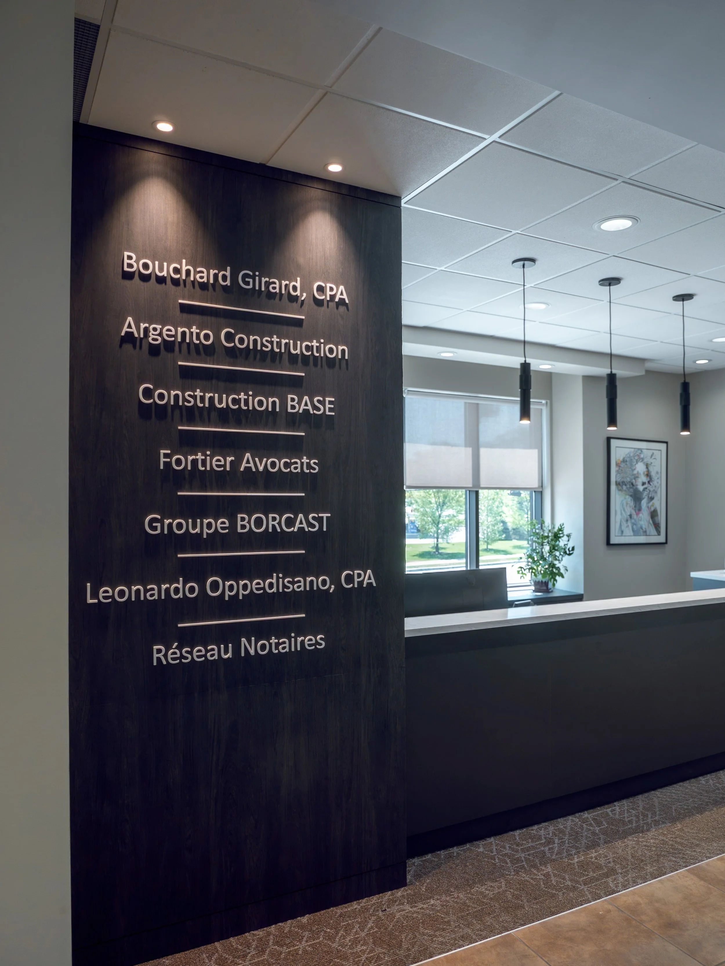 Office reception area with a black wall displaying names of professionals and firms, including Bouchard Girard, CPA, Argento Construction, Construction BASE, Fortier Avocats, Groupe BORCAST, Leonardo Oppedisano, CPA, and Réseau Notaires. The area fea
