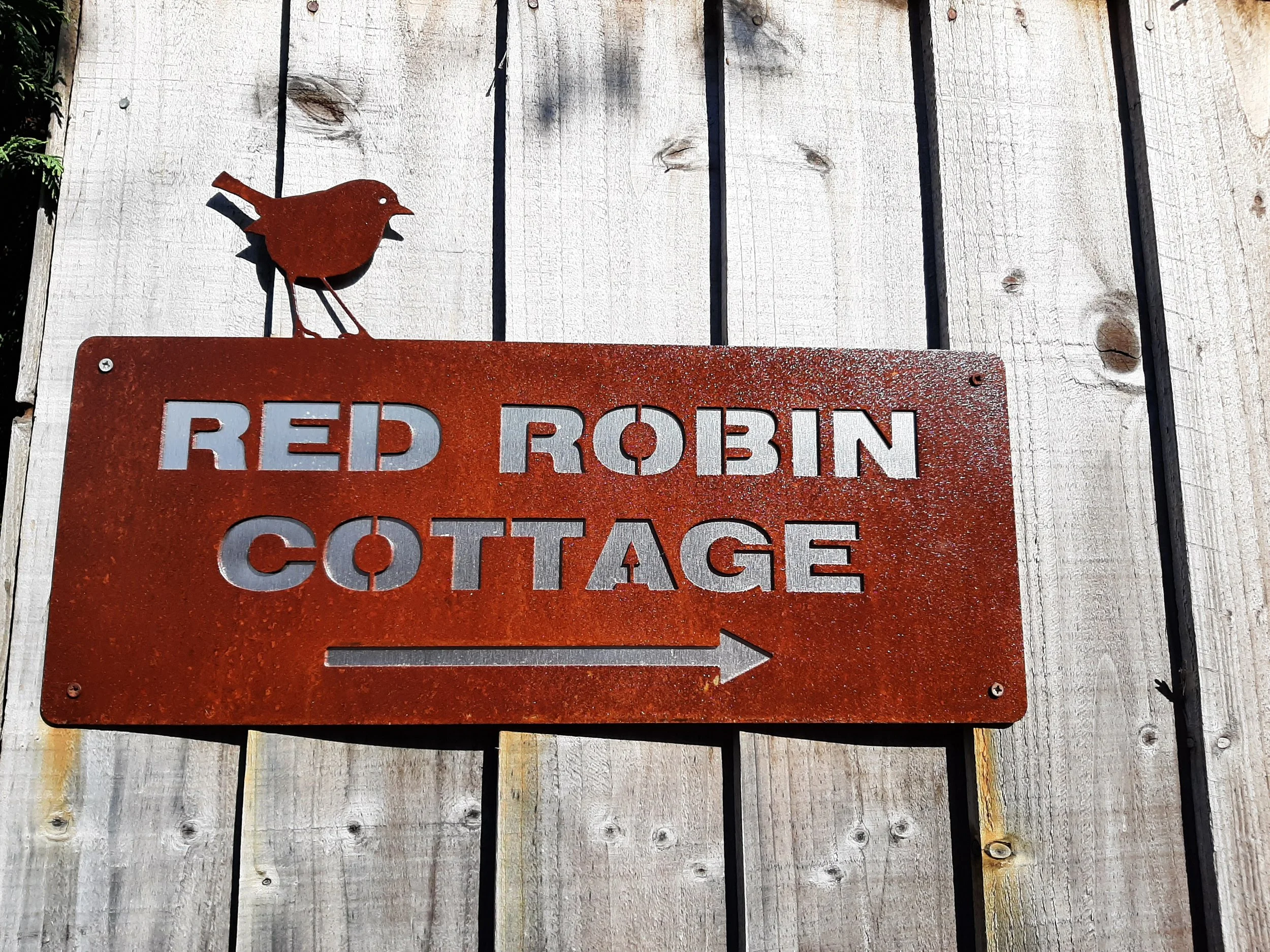 A metal sign with cut-out letters reading 'RED ROBIN COTTAGE' and a right-pointing arrow, mounted on a wooden fence with a small, rust-colored bird figure on top.