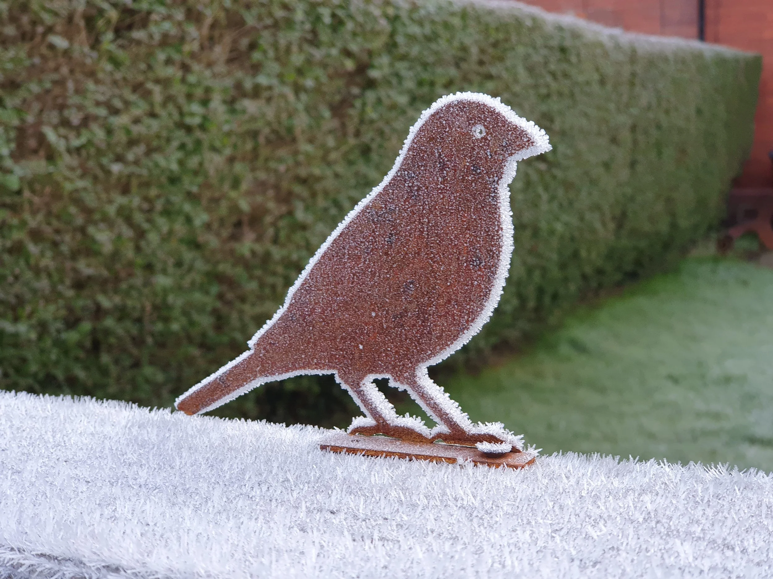 Frost-covered bird-shaped ornament on a white surface outdoors, with a background of a green hedge and red brick wall.
