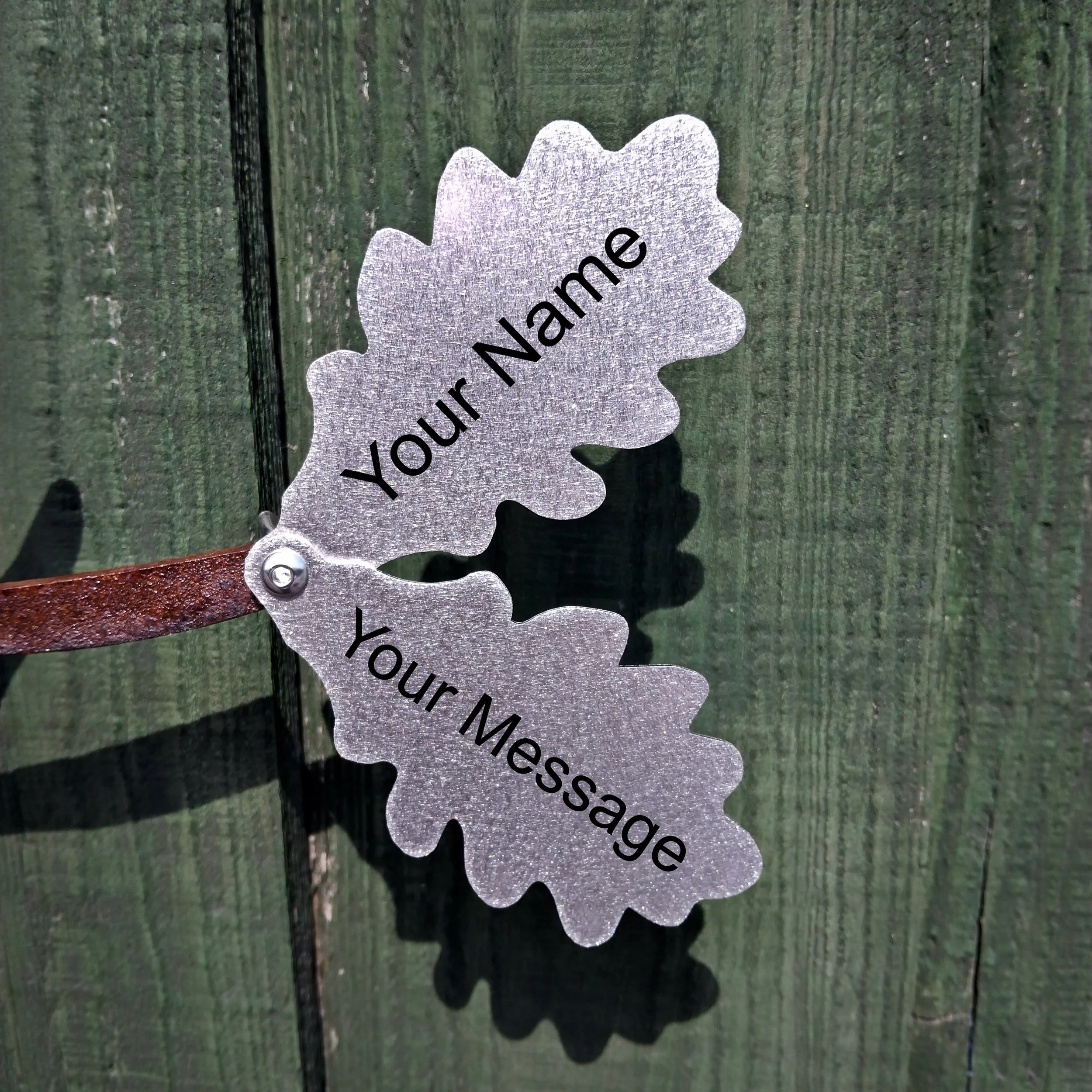 Two metallic leaf-shaped signs attached to a brown leather strap, with one sign reading 'Your Name' and the other 'Your Message', mounted on a green wooden background.