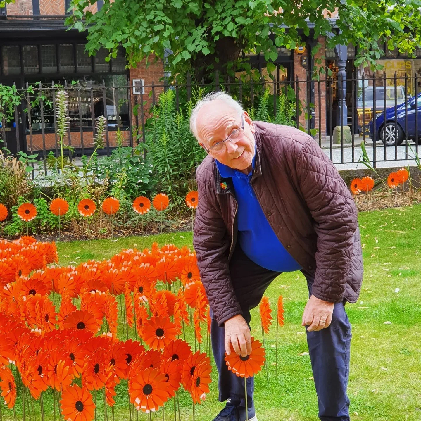 Pete waterman, is crouching and holding a bright orange metal flower among orange flowers in a garden with greenery and a black fence in the background.