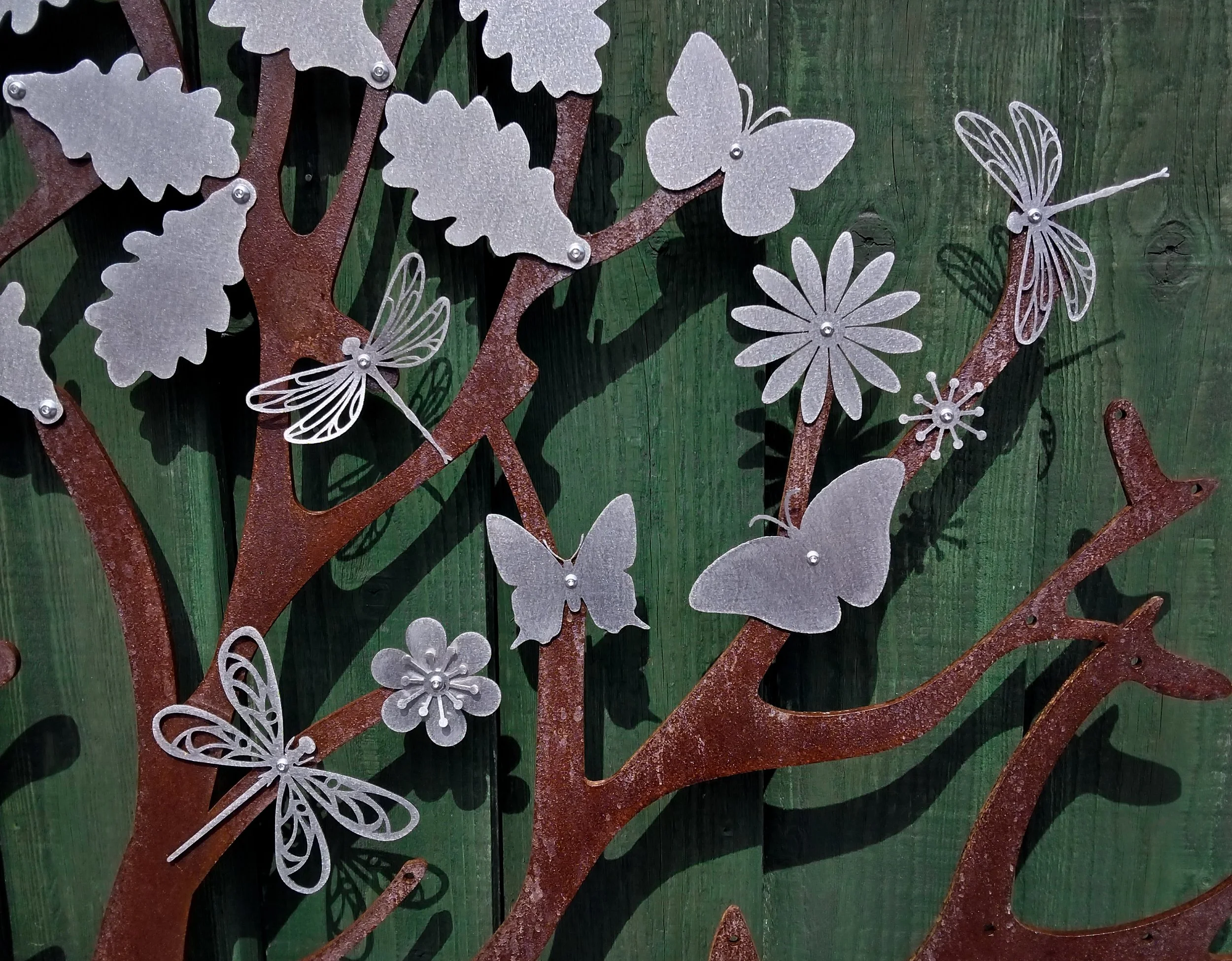 Decorative metal art of a tree with butterflies, flowers, and leaves, mounted on a green wooden background.
