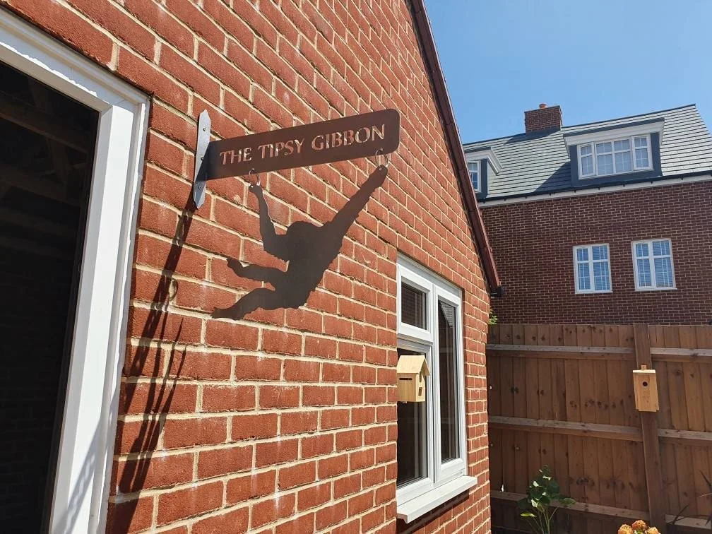 A wall-mounted sign with a silhouette of a person climbing, labeled 'The Tipsy Gibbon', casts a shadow on a brick wall in sunlight. There's a window below the sign with a small birdhouse hanging outside, and a wooden fence with a plant in the yard.