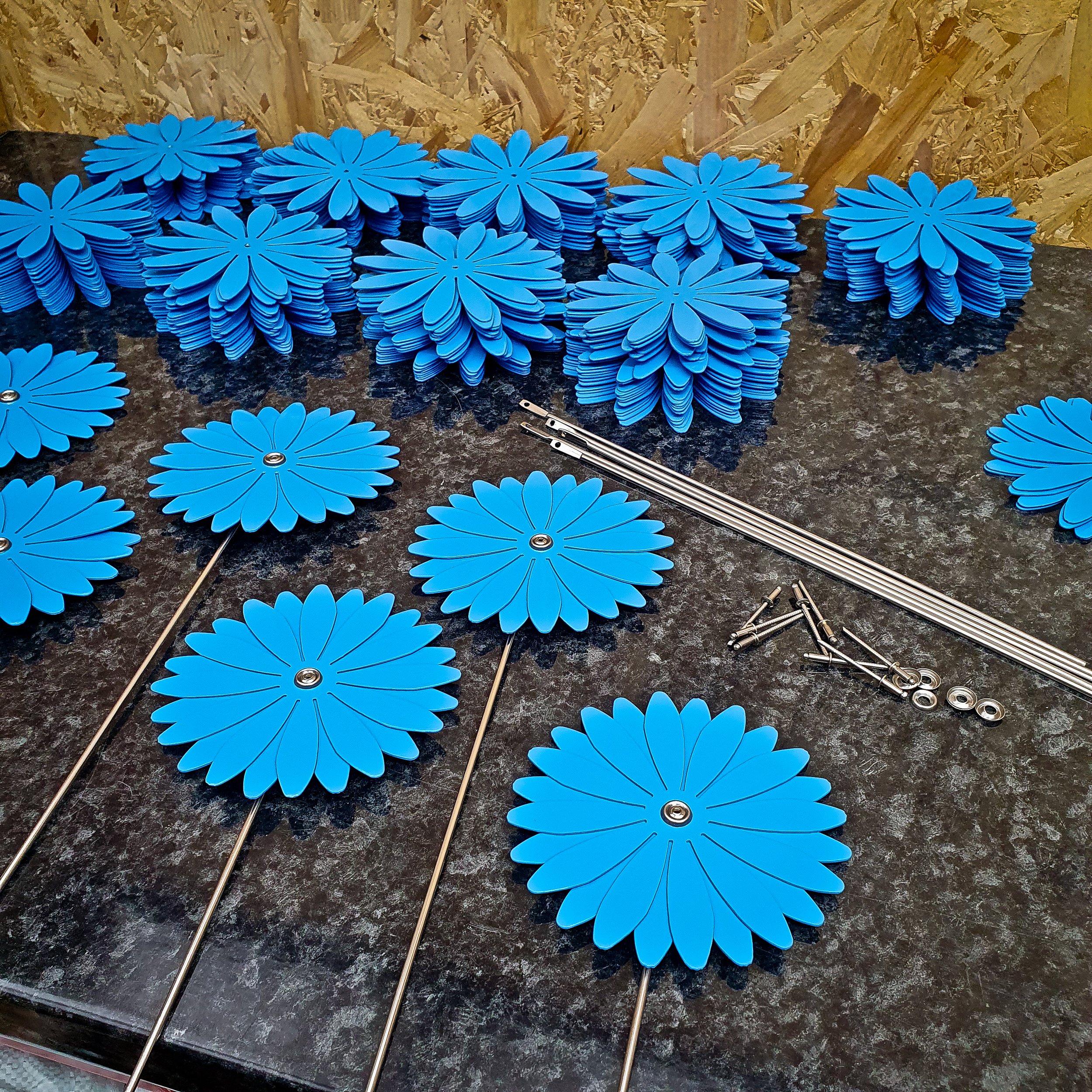 Multiple blue daisy-shaped window screens laid out on a black work surface, with some disassembled parts and metal rods nearby.