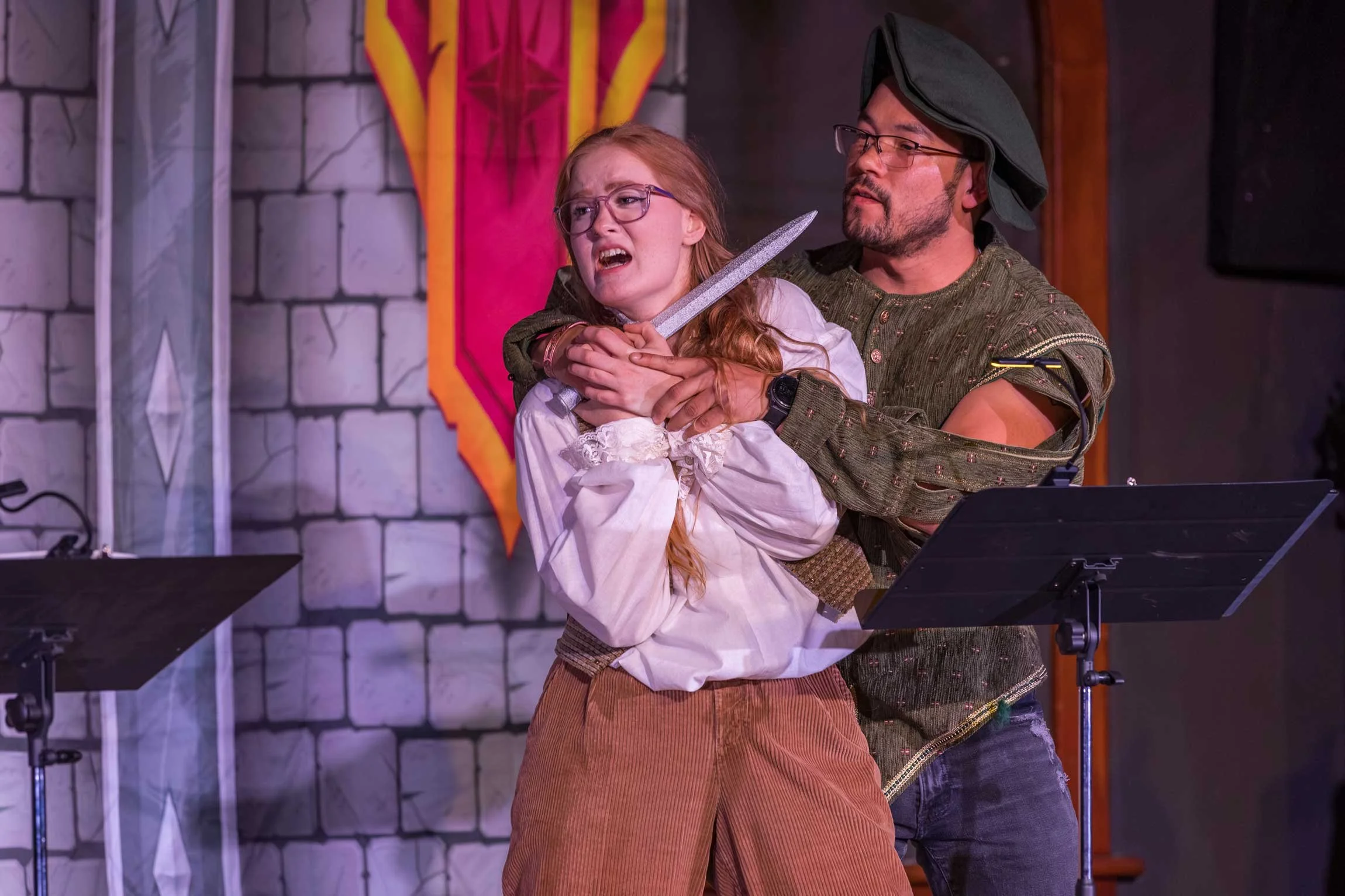 A man dressed as a knight holding a sword and a young girl dressed in period clothing with red hair, both on a stage with a stone wall backdrop and a colorful banner in the background.