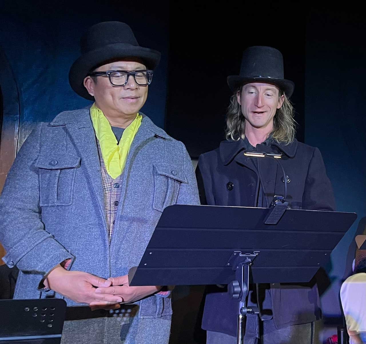 Two men wearing black hats standing at a music stand, possibly during a performance or rehearsal.