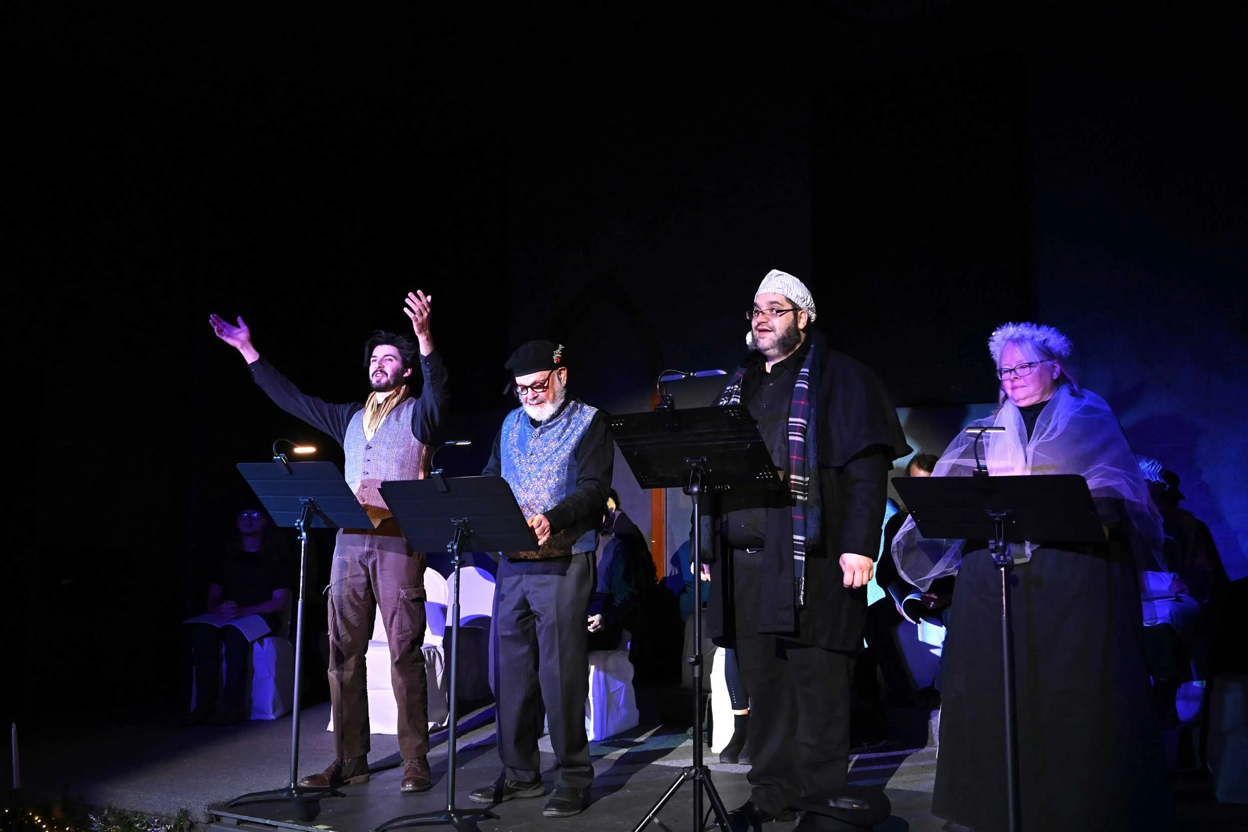 Group of four people on stage performing, dressed in festive and vintage clothing, with music stands and microphones, in a dark theater setting, some audience members seated behind them.