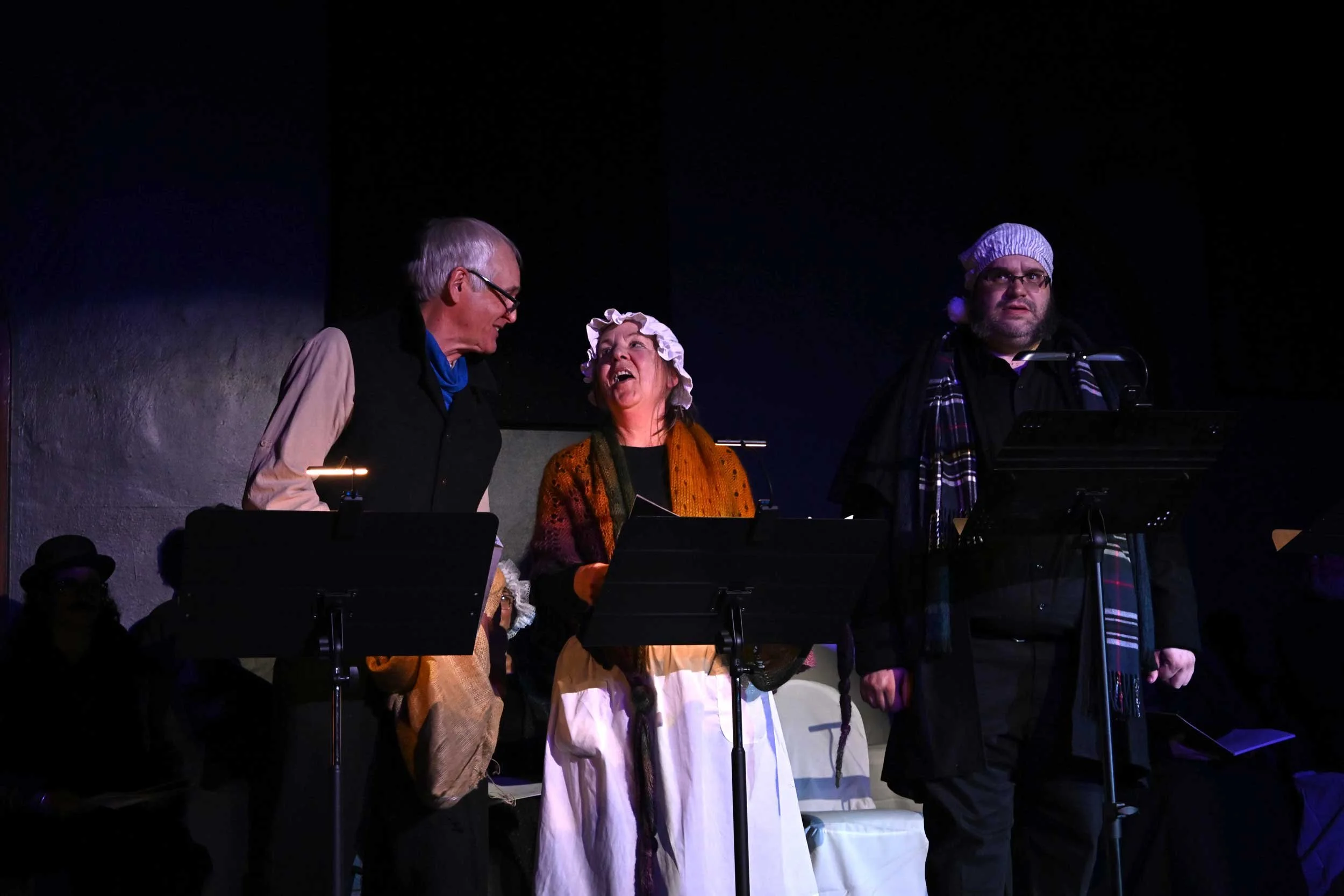 Three people standing on stage, two women and one man, engaged in a performance or conversation. The woman in the middle is smiling and wearing a bonnet and a colorful shawl. The man on the left is looking at her, wearing glasses and a vest. The person on the right, dressed in dark clothing with a scarf and a hat, appears to be focused, possibly singing or reading. Microphone stands are visible in front of them.