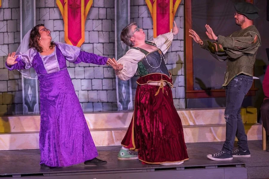 Three actors on stage performing in a theatrical play with medieval costumes, set against a castle backdrop.