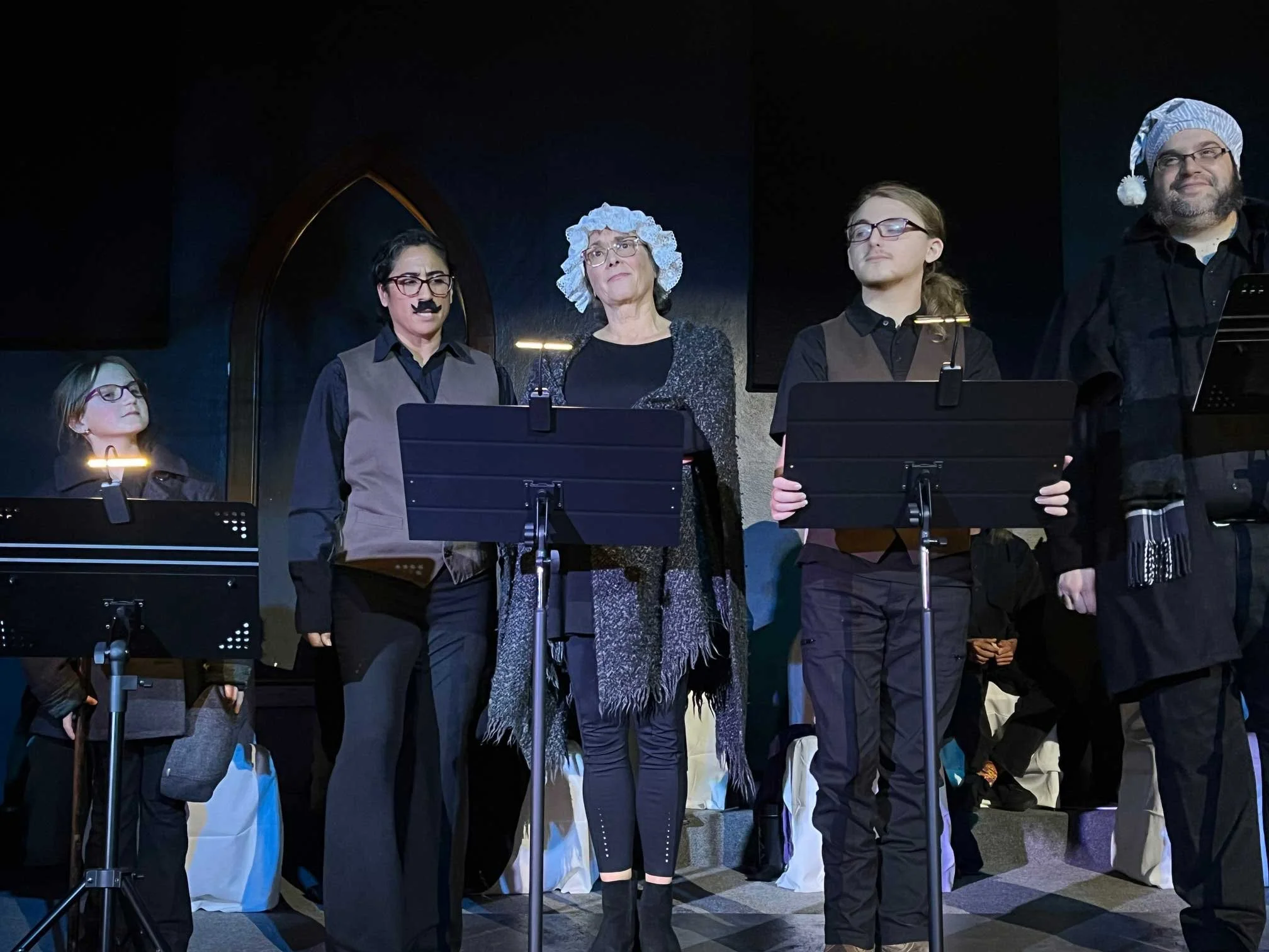 Group of five people dressed as a choir performing on stage, some wearing costumes and props, with music stands in front of them, against a dark background.