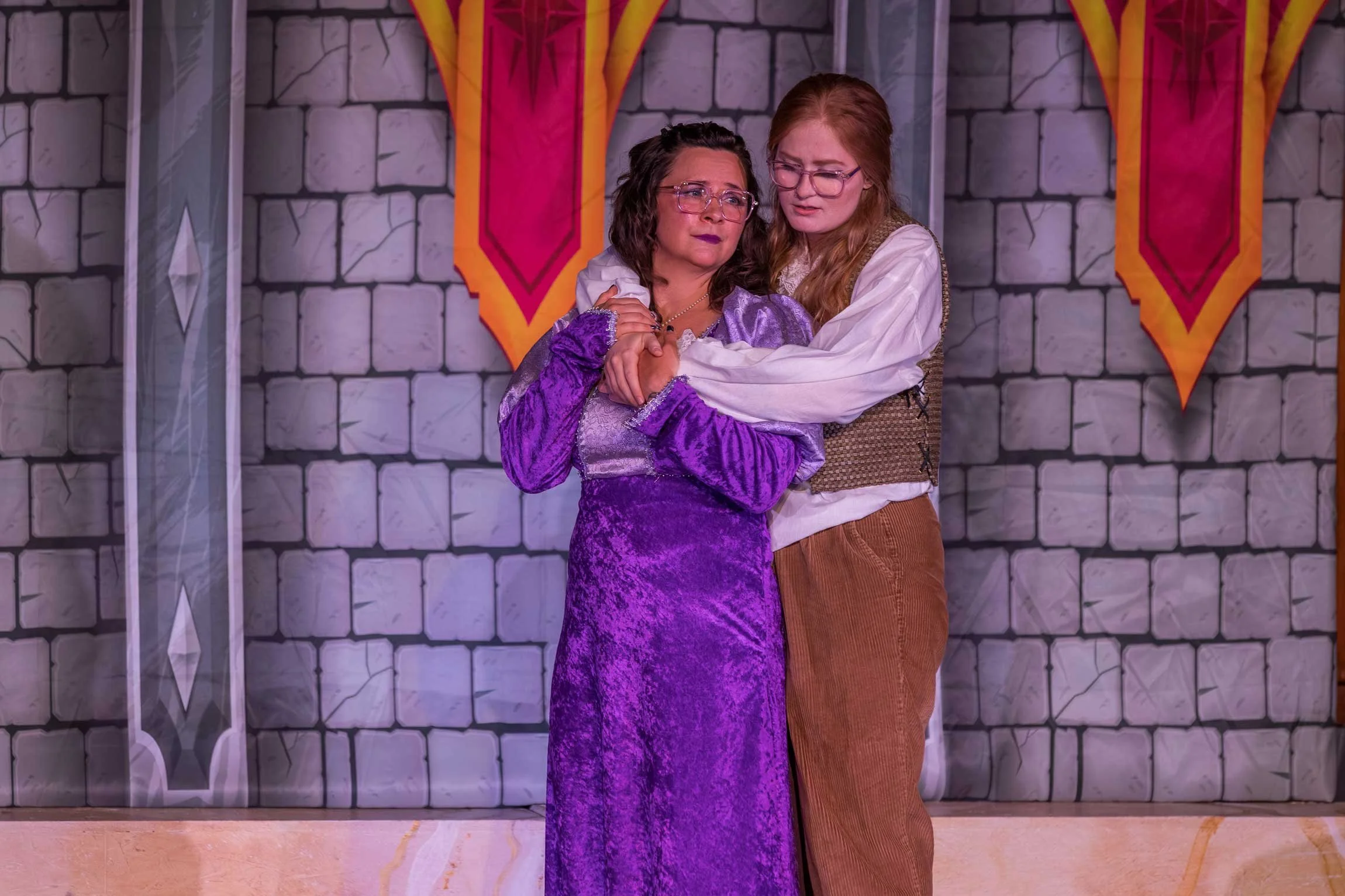Two women, one in a purple dress and the other in brown pants and a white shirt, embrace each other on a stage with a castle-like stone wall backdrop and castle banners.