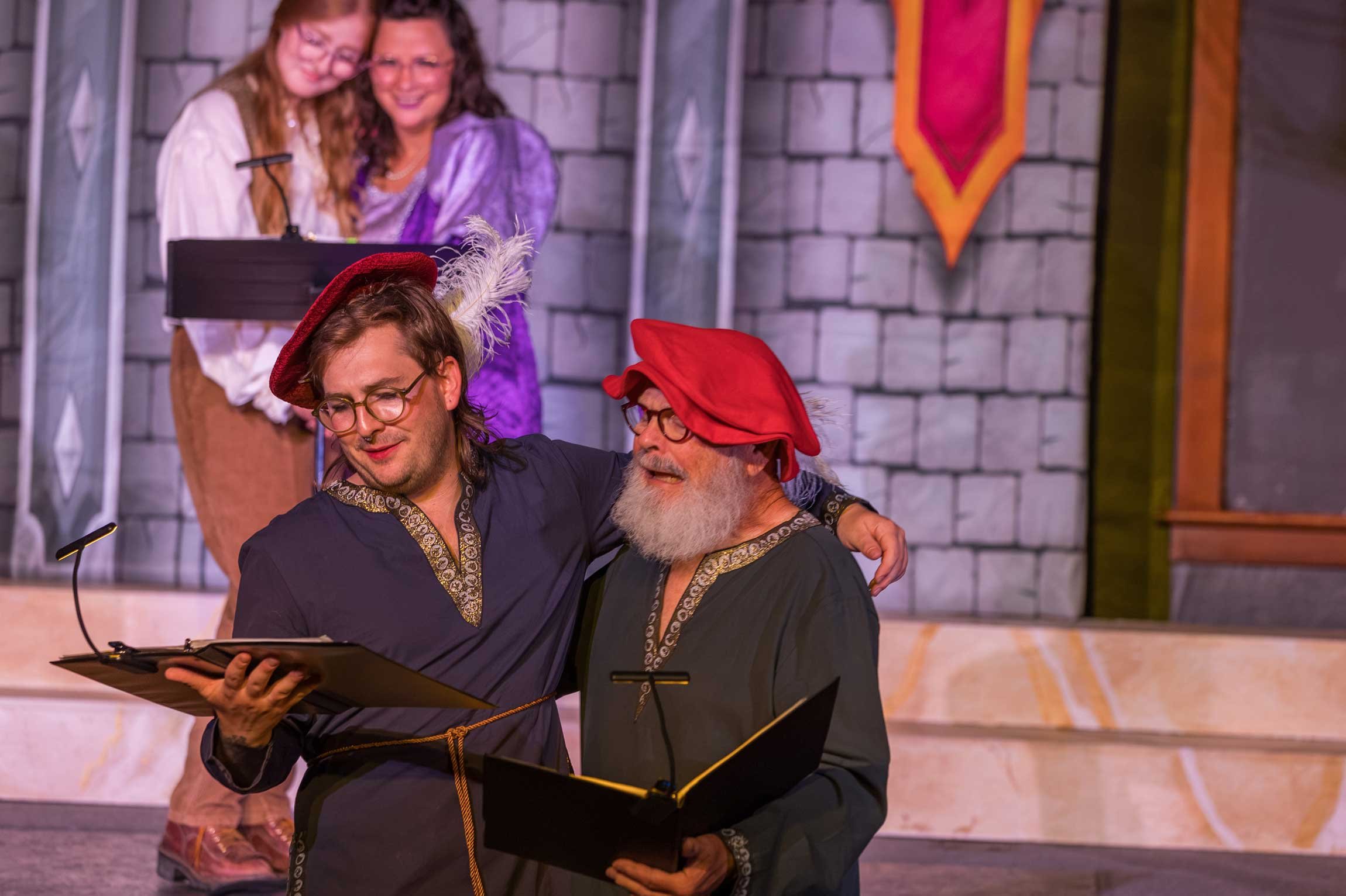 Two men dressed as wizards with red hats and robes, reading a book together, standing close to each other with one man's hand on the other's shoulder, on a stage with a castle wall backdrop, with two women in medieval costumes in the background.