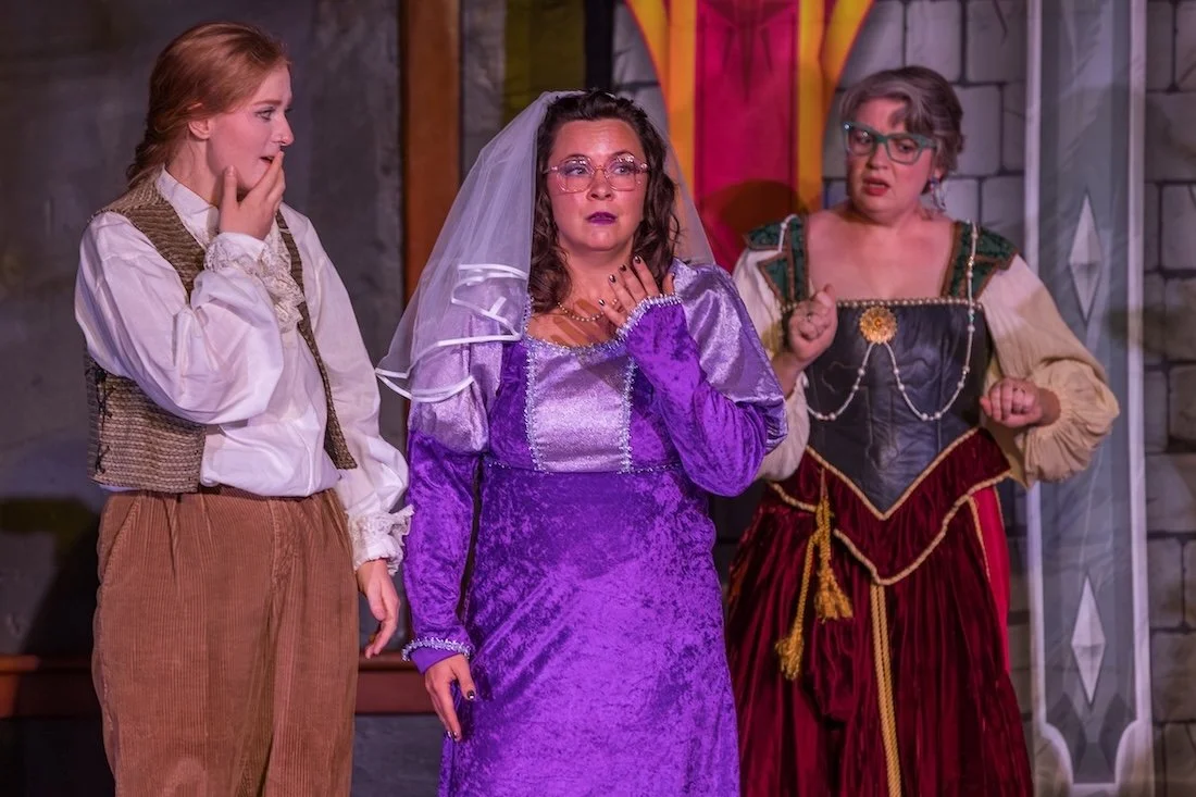 Three women in costumes on stage, with medieval castle background, performing in a play.