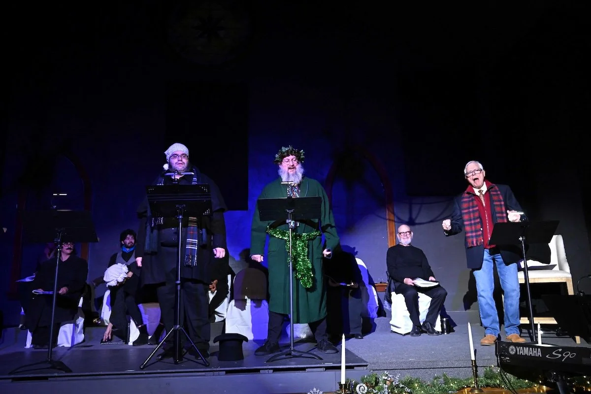 A group of elderly men performing on stage, dressed festively with one wearing a Santa hat, another with a holiday wreath, and another in a red plaid jacket, surrounded by Christmas decorations.