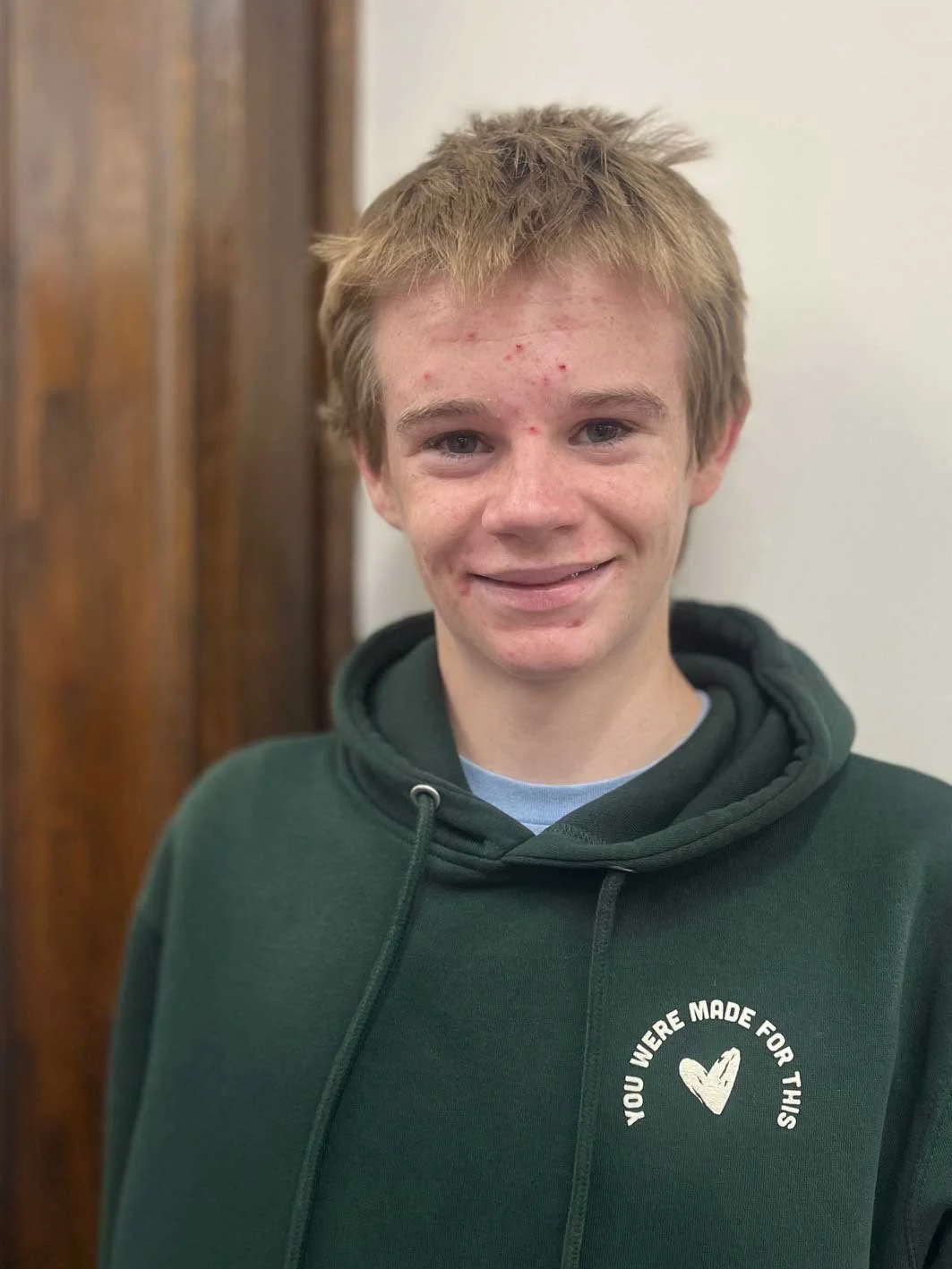 Young person with short blond hair and acne, wearing a dark green hoodie with a heart graphic and text, smiling indoors near a wooden wall.