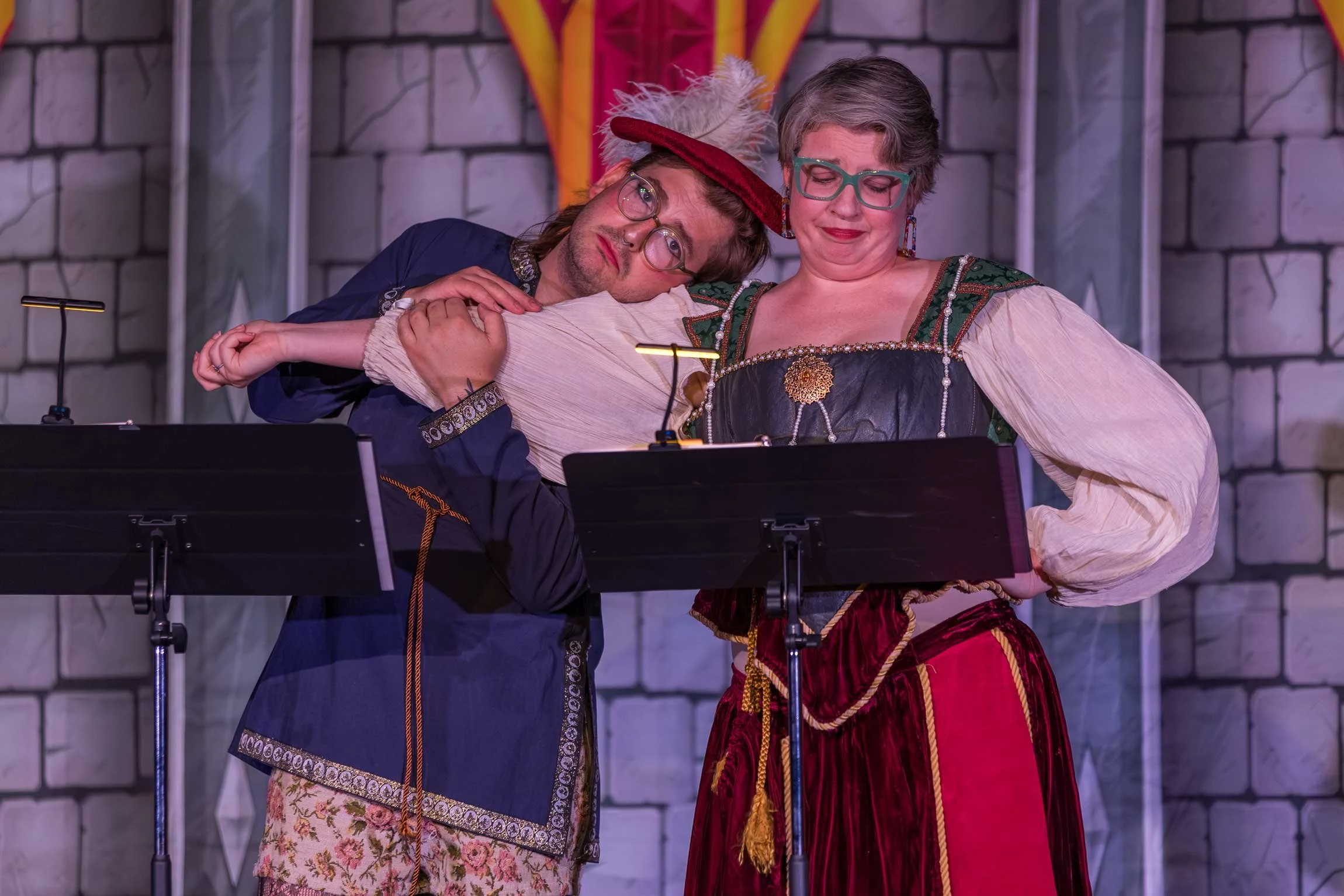 Two individuals dressed in renaissance costumes, one appearing to be asleep or resting on the other's shoulder. They are standing behind music stands, against a stone wall background, in a setting resembling a theatrical performance or reenactment.