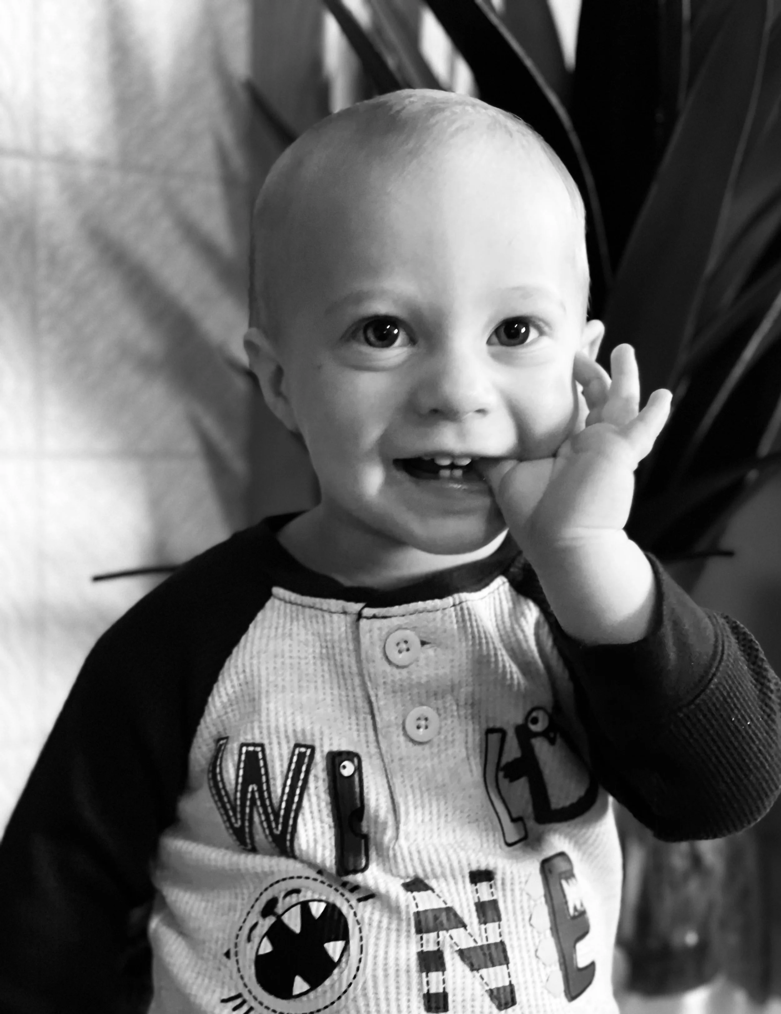 A young child with short hair, smiling and touching their face with one hand, wearing a shirt with letters and graphic designs, standing indoors near a plant.