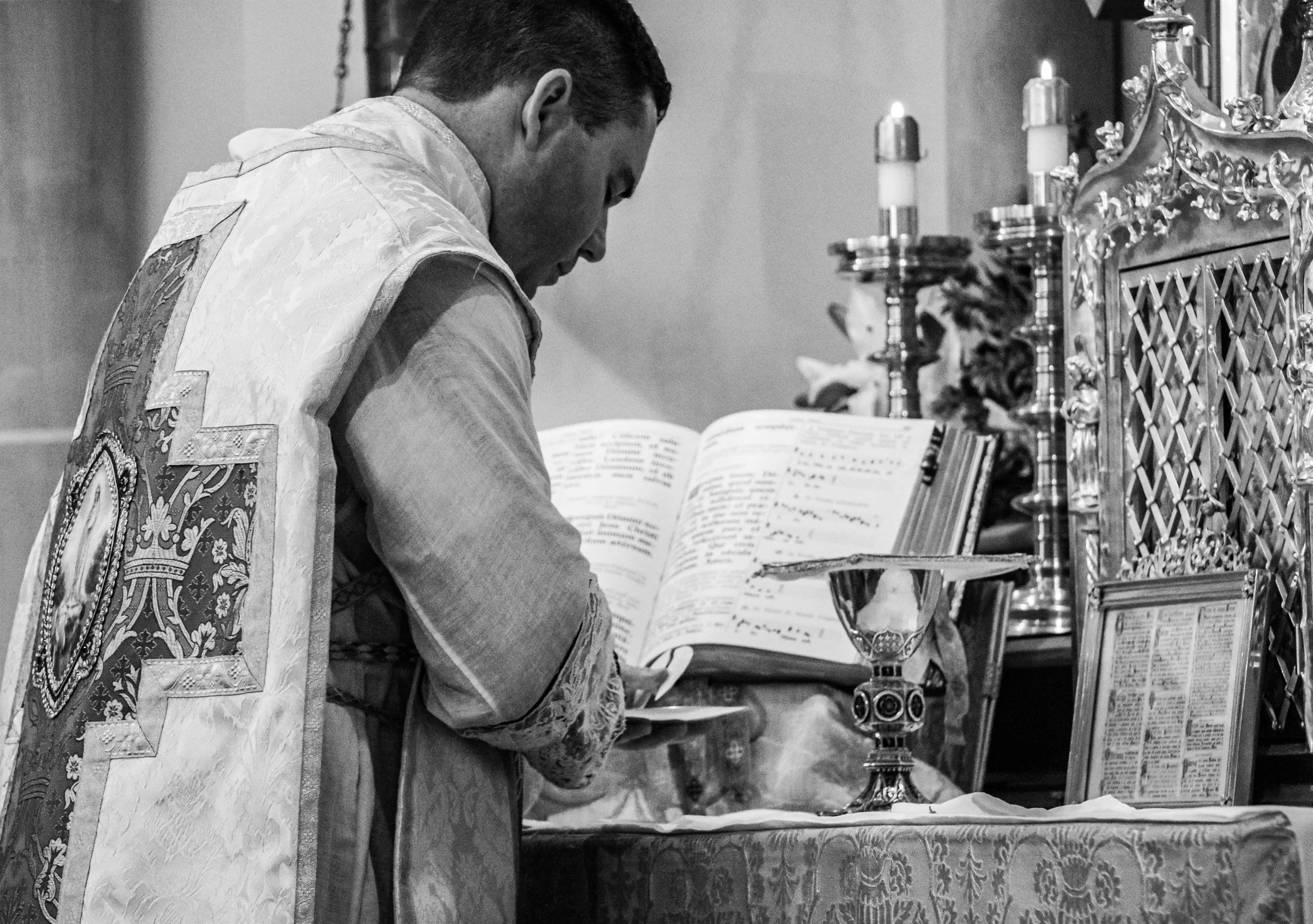 A priest in religious vestments performing a sacred ritual at an altar with lit candles, an open liturgical book, and ornate religious items in a church setting.