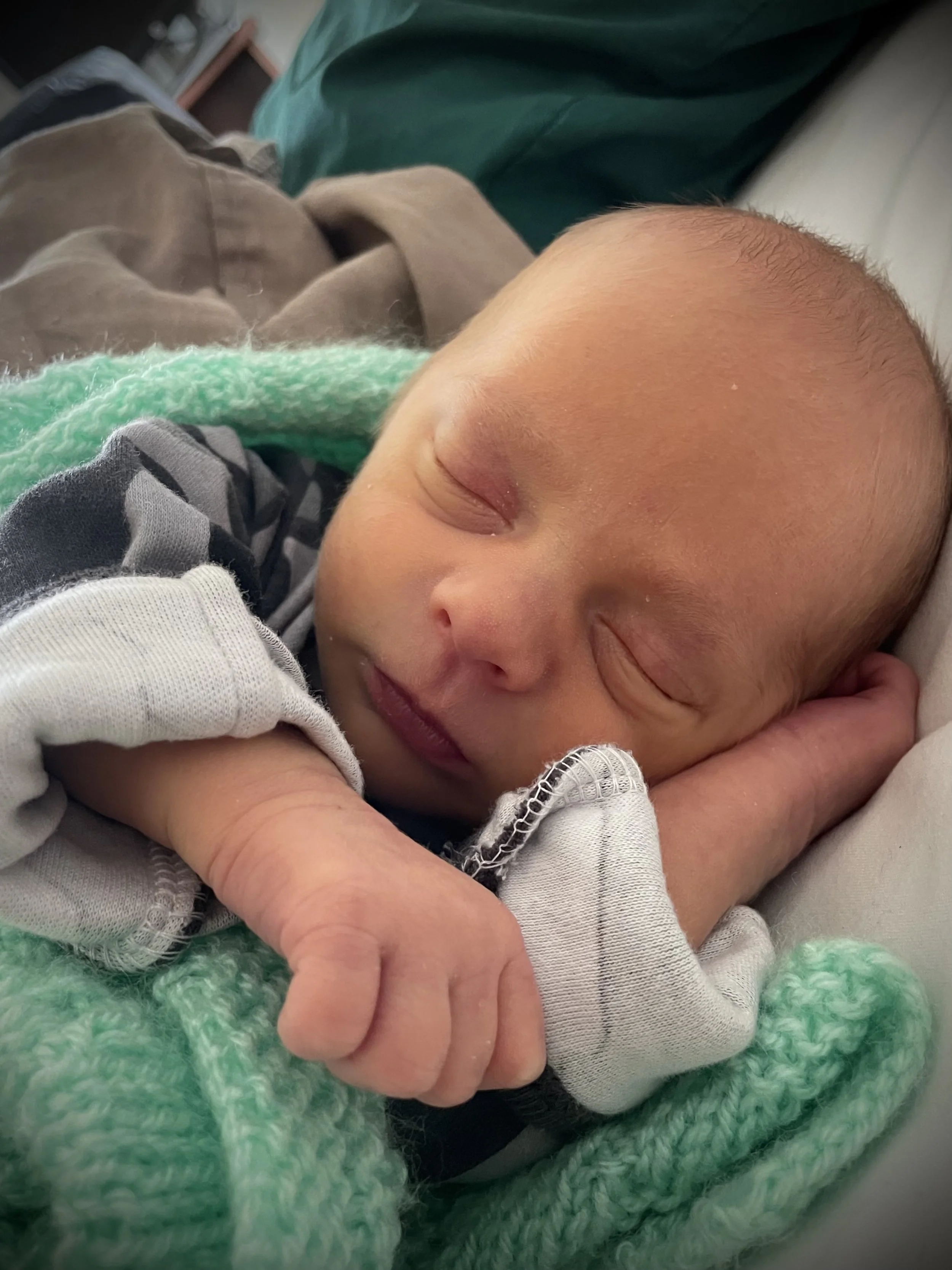 A sleeping newborn baby with closed eyes, lying on a person's arm, wrapped in a green knitted blanket.