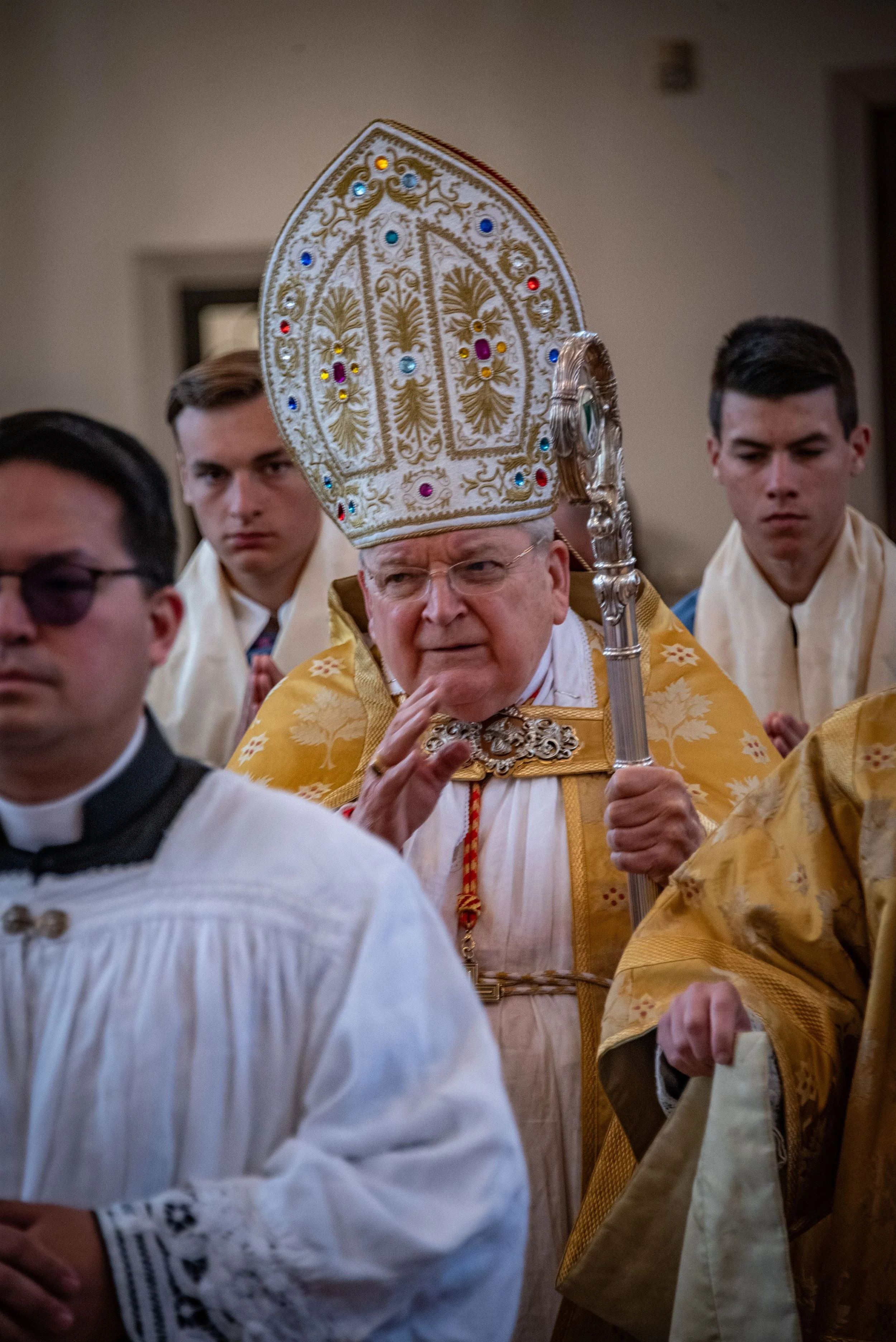 An elderly man dressed as a bishop or pope holding a staff, wearing an ornate mitre, participating in a religious ceremony surrounded by young men in robes.