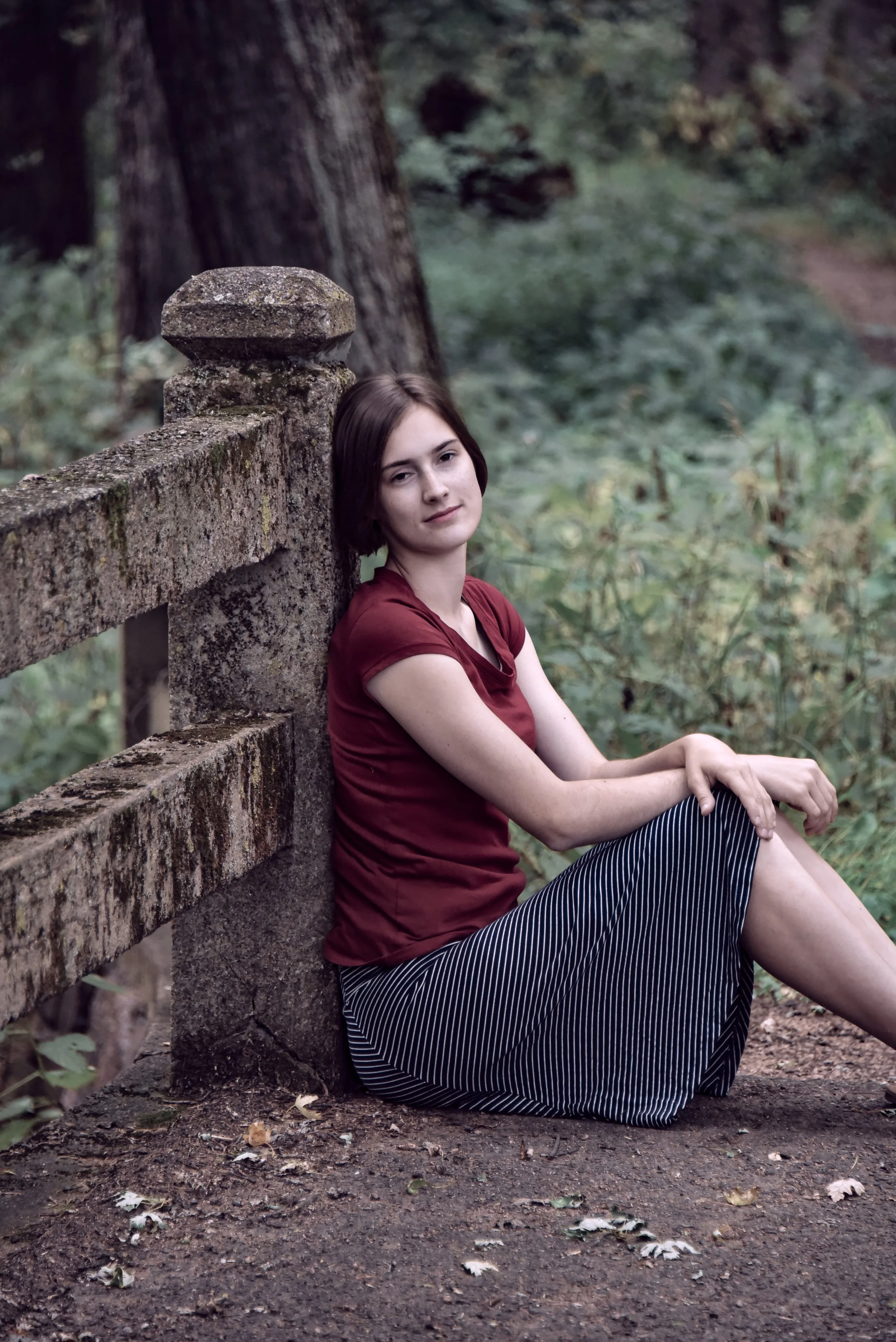 A young woman with short dark hair sitting on the ground next to a weathered stone wall in a forest, wearing a red t-shirt and a black-and-white striped skirt.