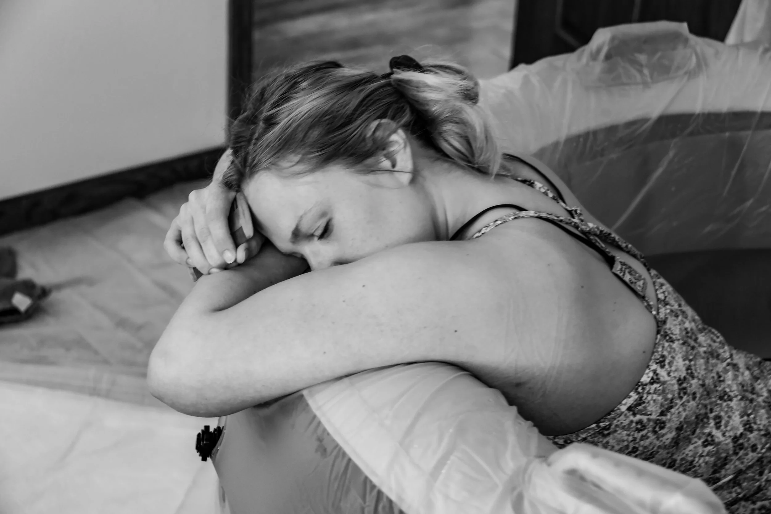 Black and white photo of a woman with closed eyes resting her head on her arm as she labors in a birth pool, appearing peaceful but tired.