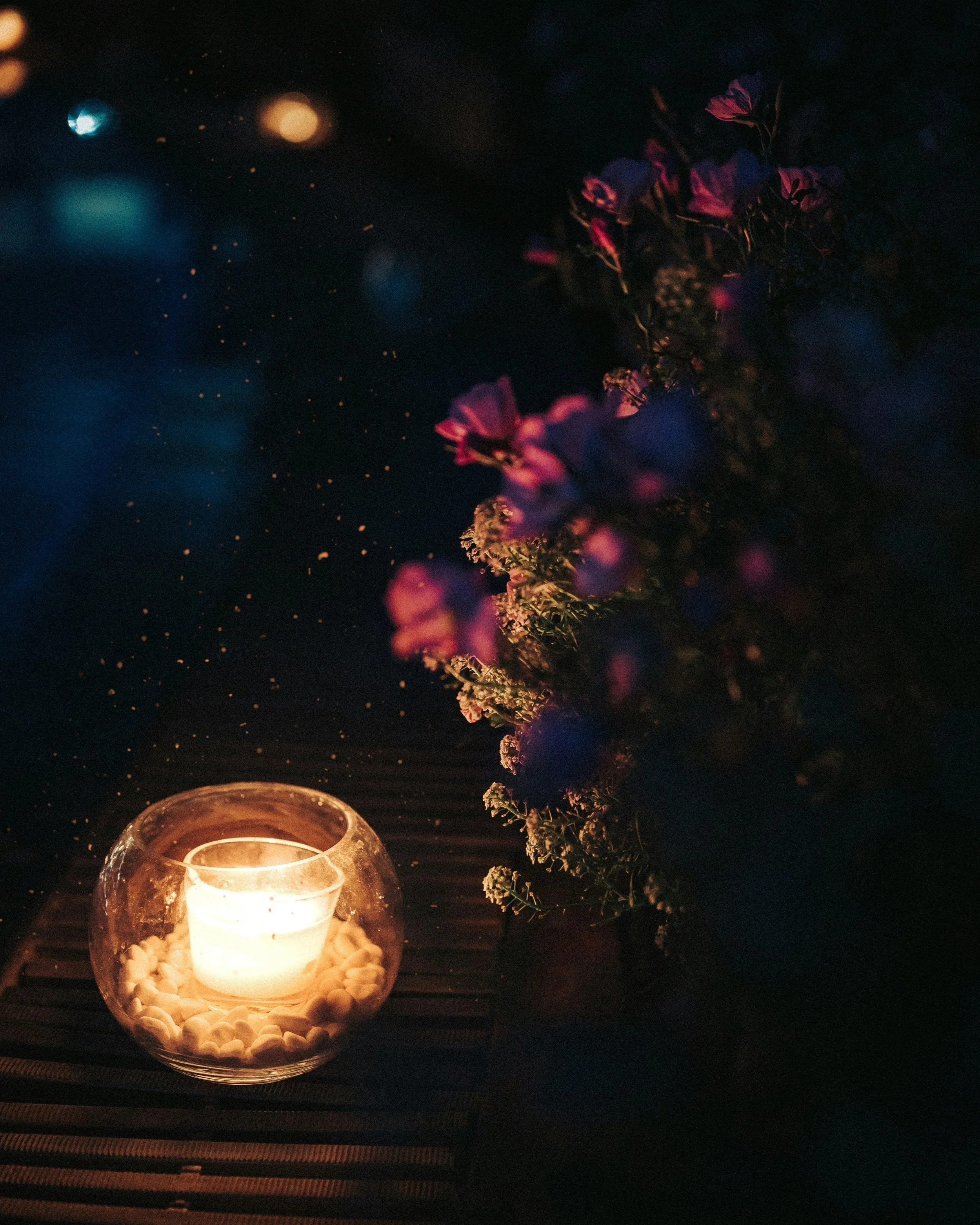 A lit candle in a glass holder surrounded by small white pebbles, placed on a dark wooden surface, with a bouquet of purple flowers in a vase nearby in a dark setting.