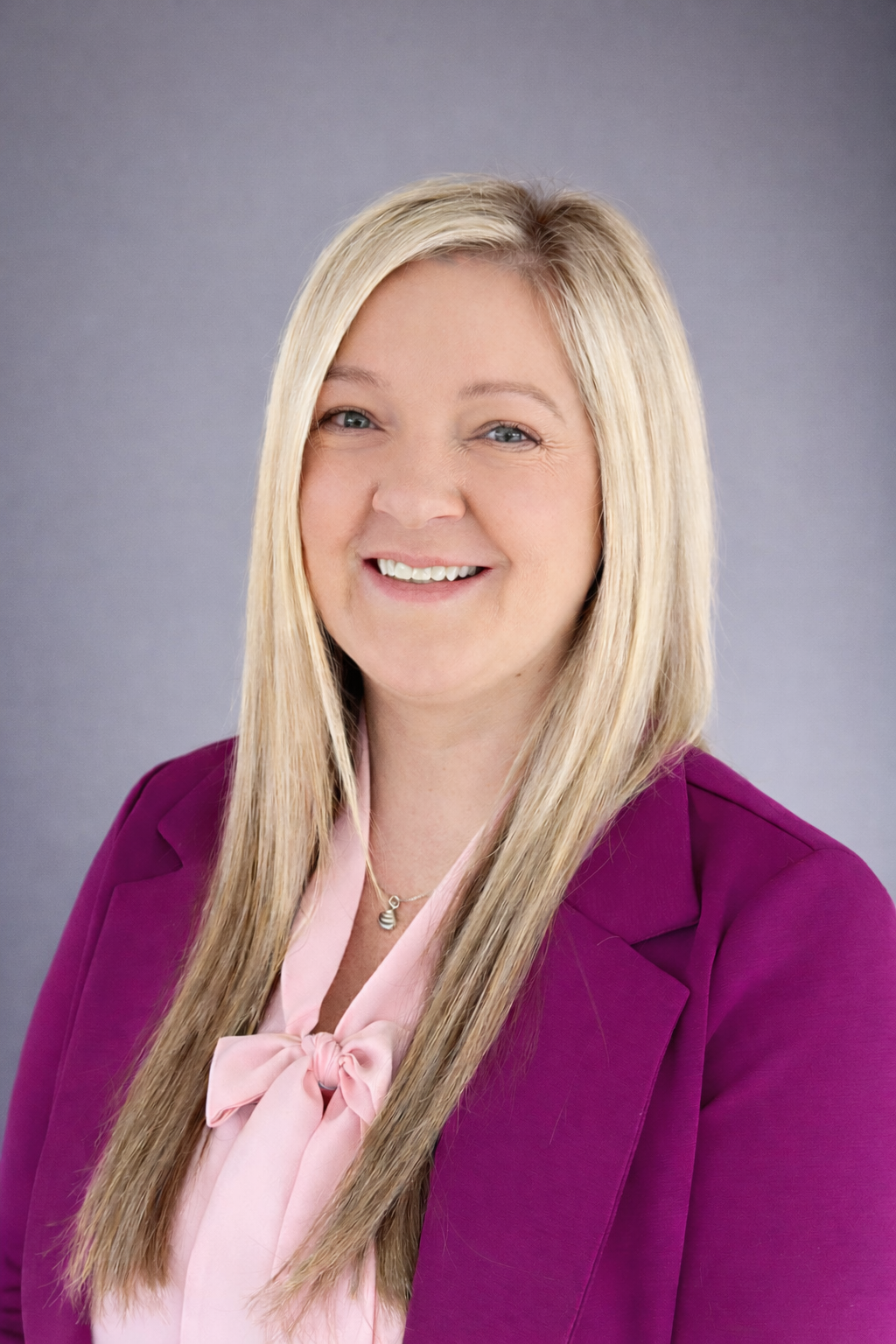 A smiling woman with long blonde hair, wearing a pink blouse and a magenta blazer, posing against a gray background.