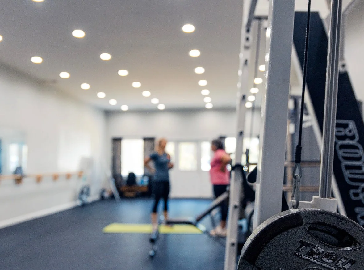 Two women in a gym conversing, with exercise equipment and a ladder in the background, and a weight plate in the foreground.