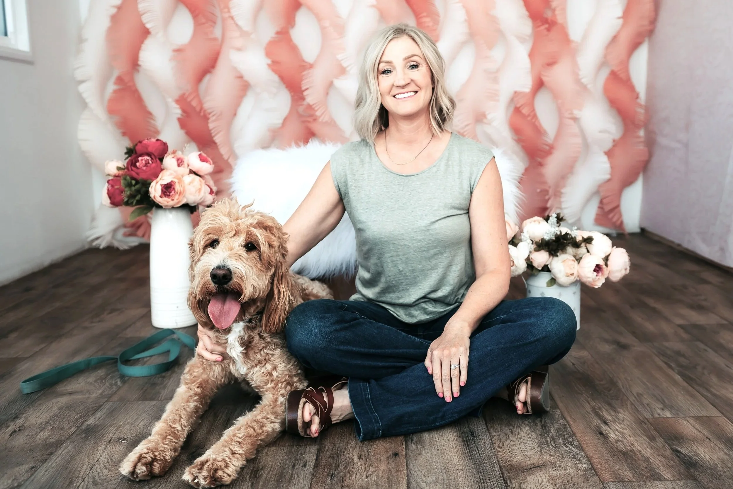 A woman with short blonde hair sits on the floor with a curly-haired dog on her lap. She is smiling and wearing a light green sleeveless top and dark blue jeans. Behind them is a pink and white feathered backdrop and two white vases with pink and white flowers. The woman is sitting on a wooden floor.