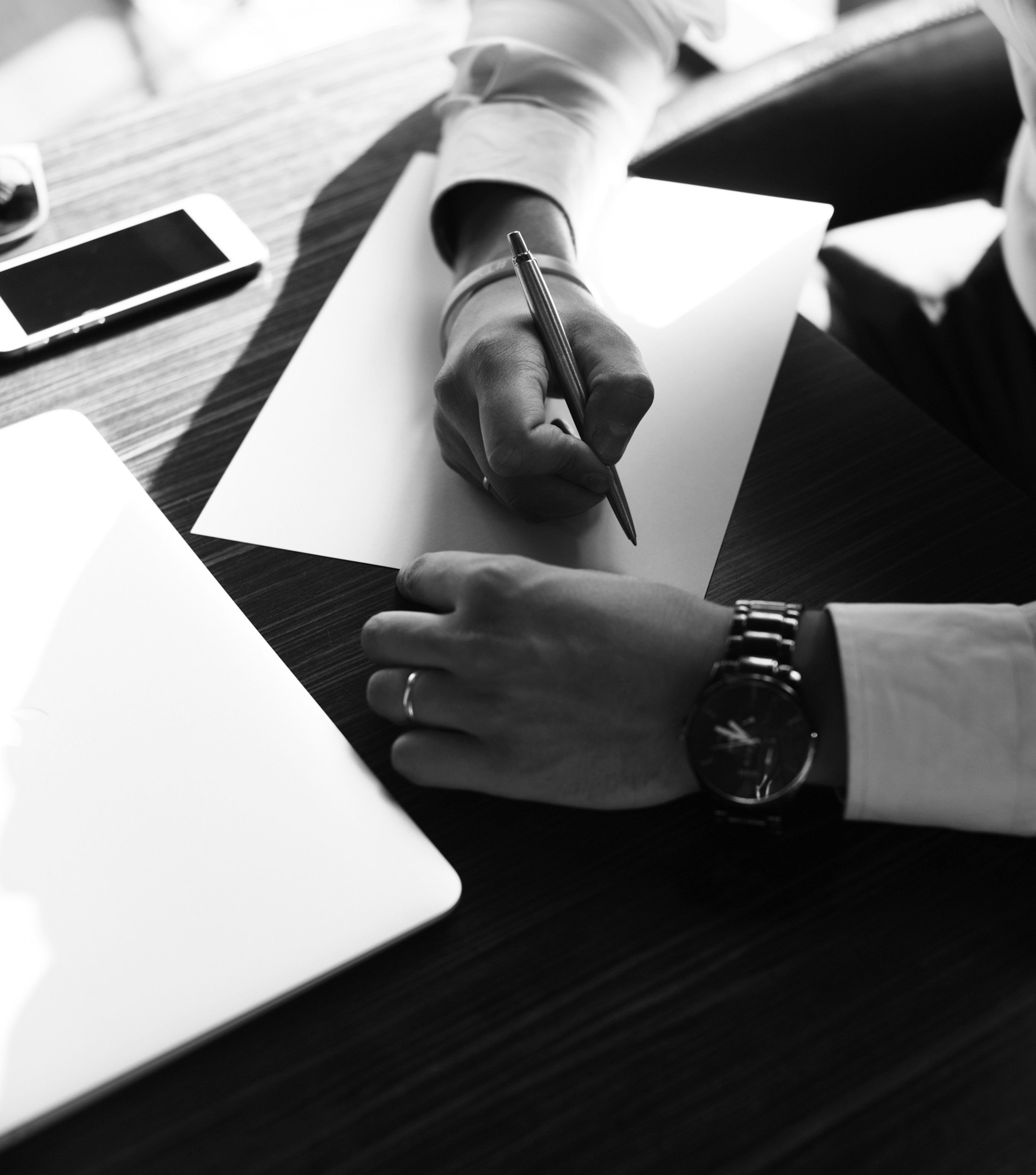 Black and white photo of a man's hands with white shirt cuffs showing holding a pen about to write on a blank piece of white paper.