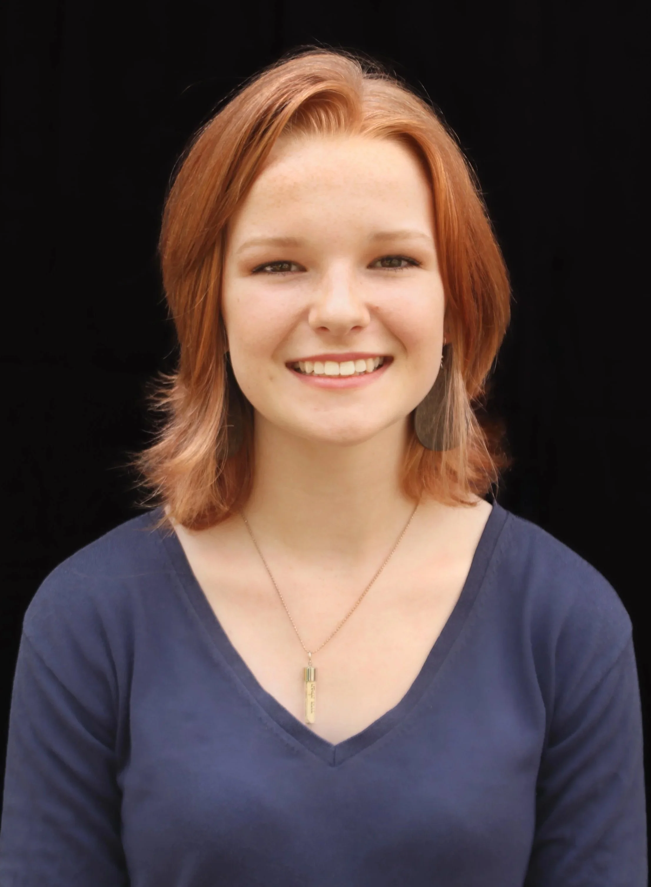 Young woman, Liberty Snyder, with shoulder-length red hair, wearing a navy blue V-neck shirt and a gold pendant necklace, with a genuine smile in front of a dark background.