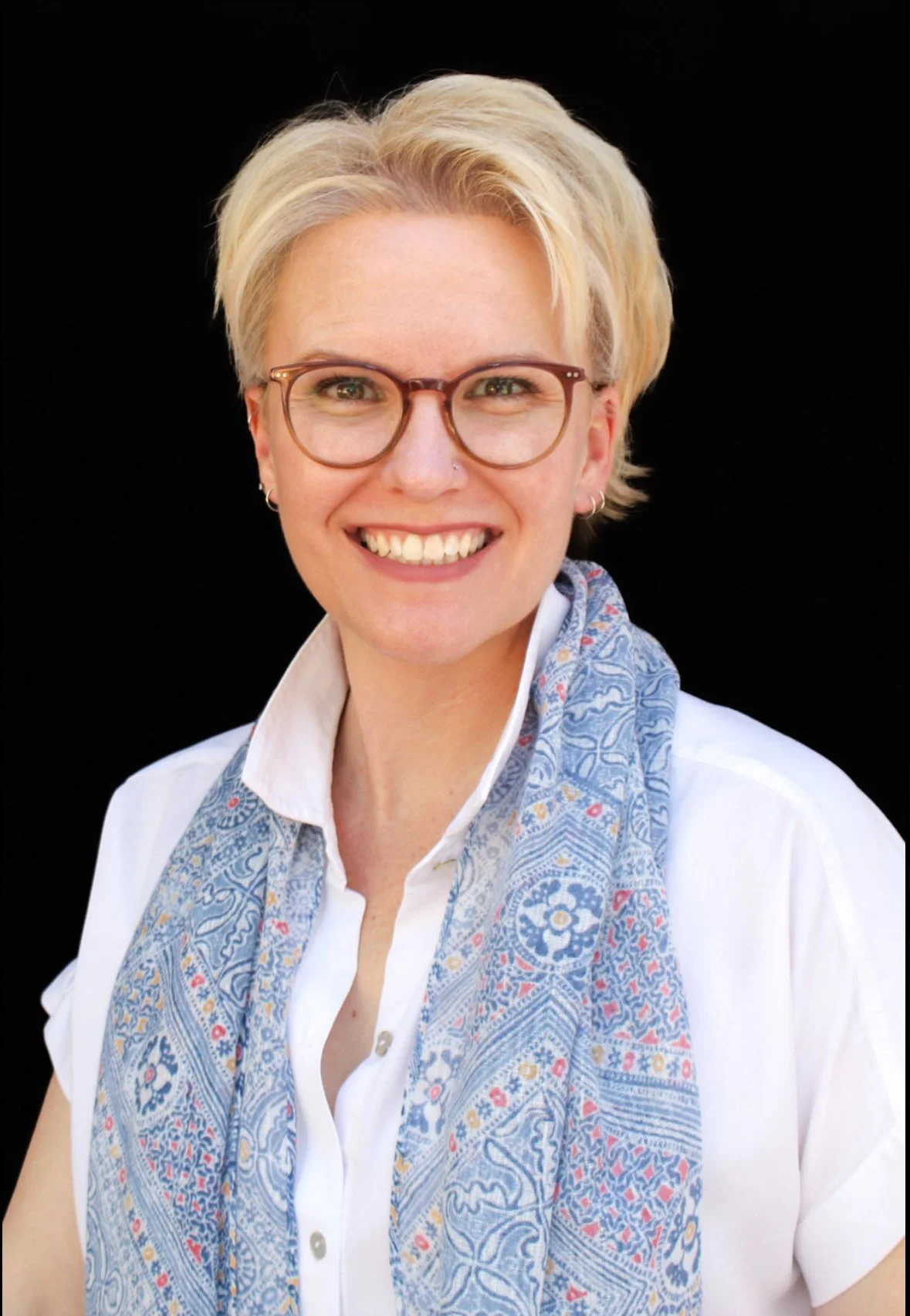 Portrait of a woman, Haley Snyder, with short blonde hair, glasses, wearing a white shirt and a blue patterned scarf, smiling against a black background.