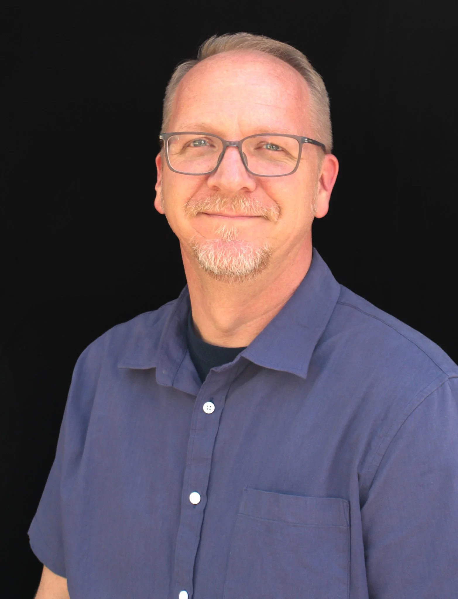 A middle-aged man, Josh Snyder, with glasses, light hair, and a beard, wearing a blue button-up shirt, smiling in front of a black background.