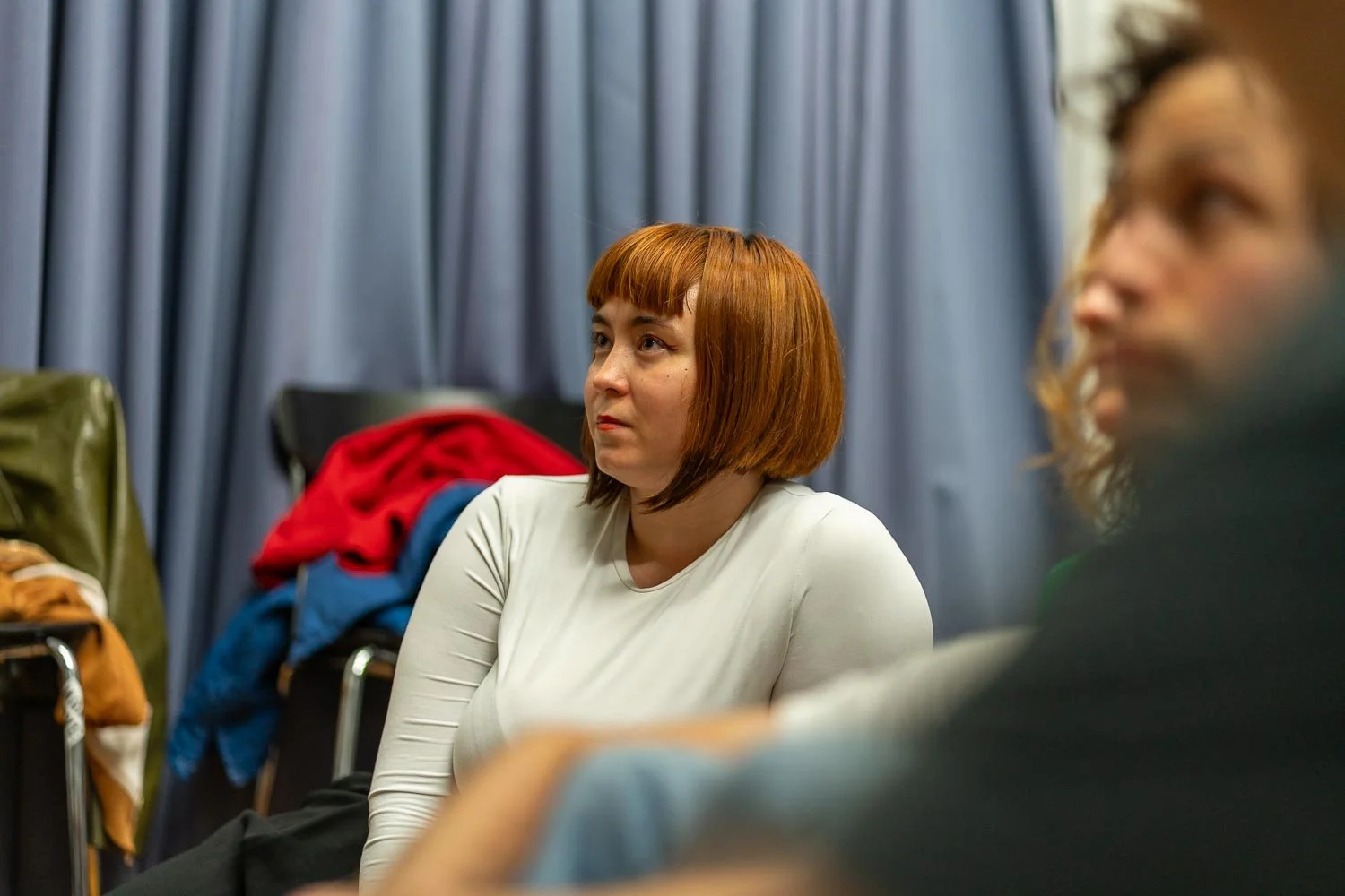 A woman with red hair in a bob haircut wearing a white top, sitting in a room with blue curtains in the background, appears to be listening attentively.