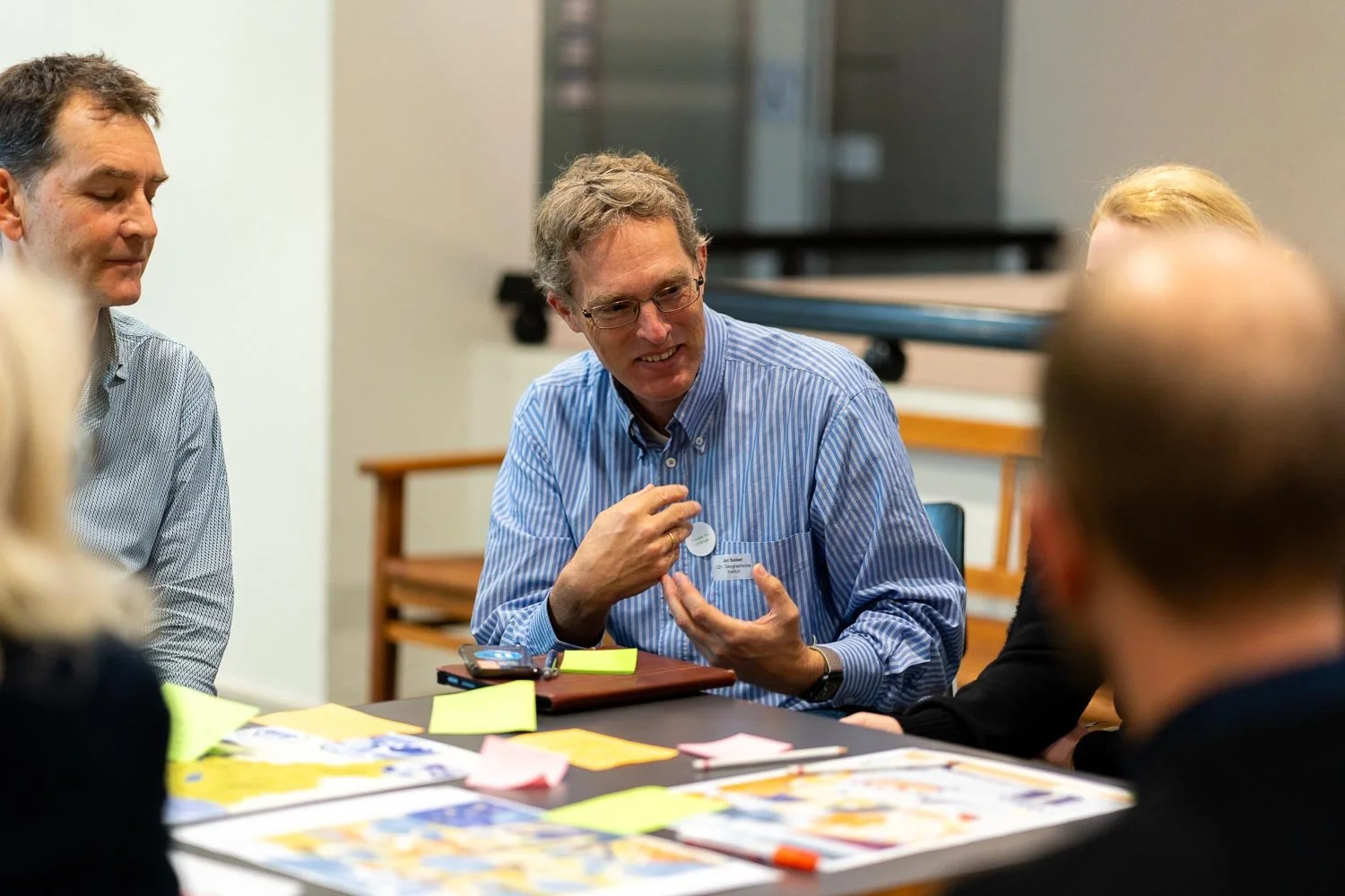 Group of professionals having a discussion around a table with papers and sticky notes, one man is explaining something with a smile.
