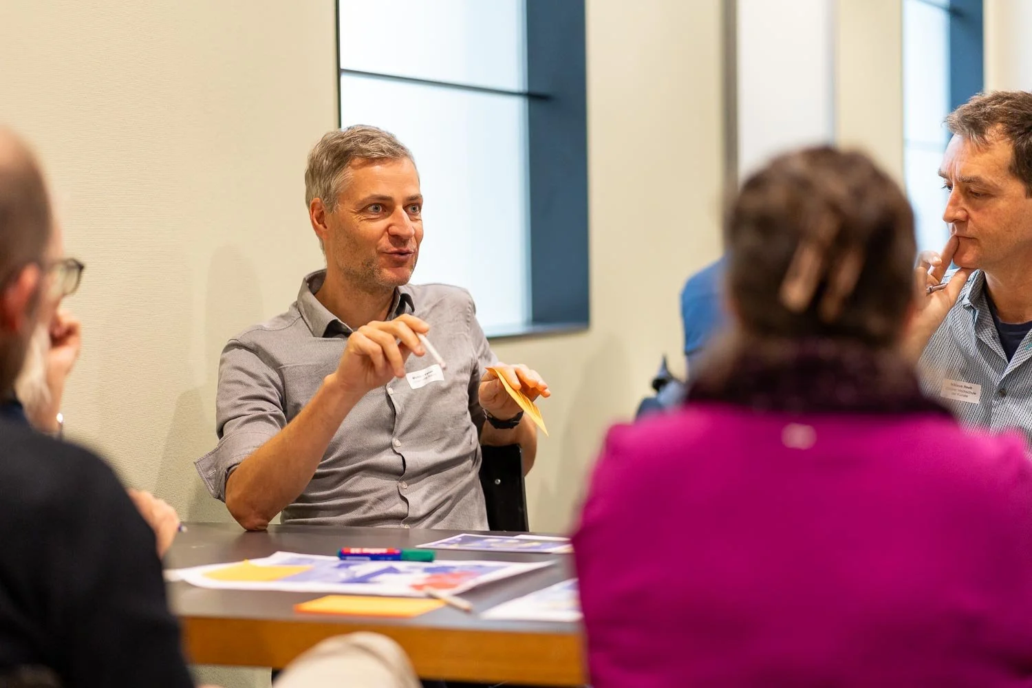 A group of five adults sitting around a table in a conference room, engaged in a discussion. The man in the center is talking and gesturing with his hands, while the others listen attentively. There are papers, markers, and a yellow sticky note on th