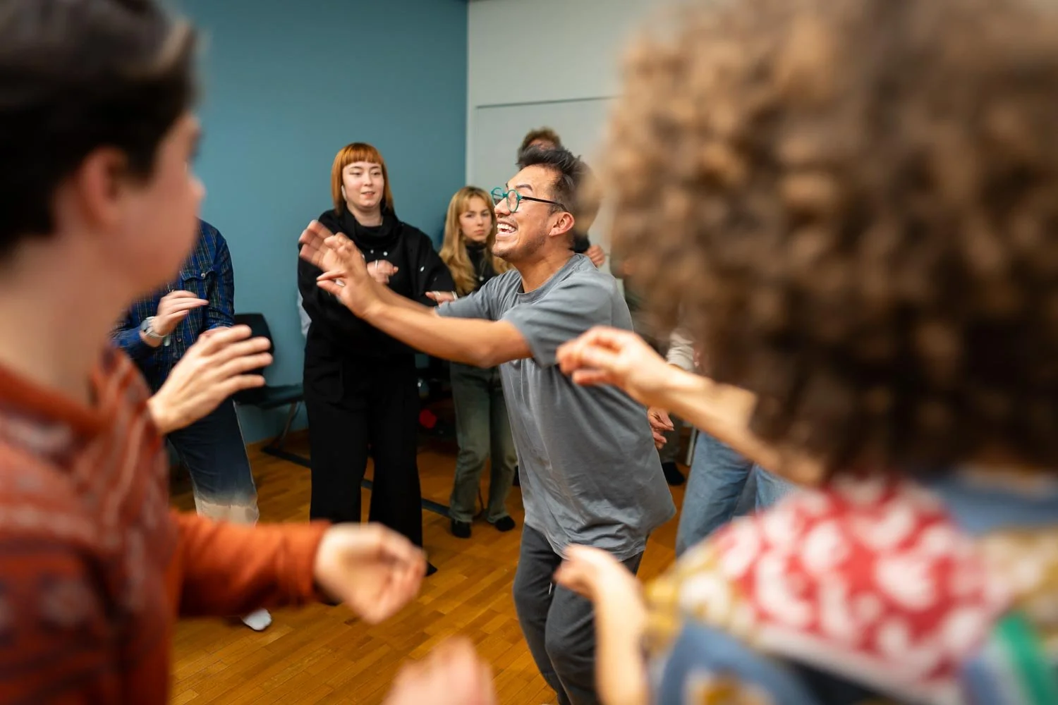 A group of people dancing and celebrating at a party or social gathering, with a man in a gray shirt laughing while interacting with others.