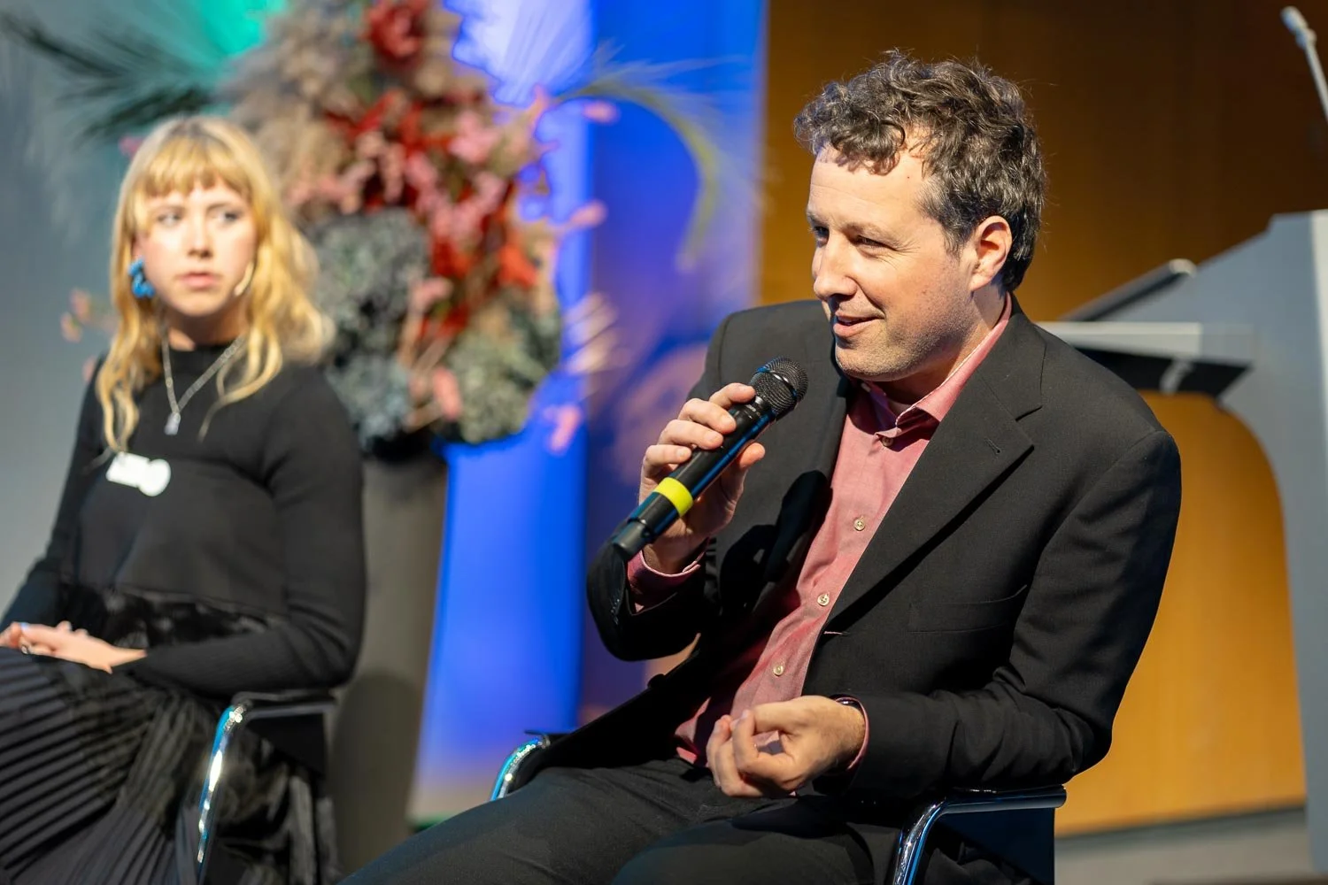 A man in a black blazer and pink shirt speaking into a microphone during a panel discussion, with a woman in black listening attentively in the background, and a floral arrangement behind them.