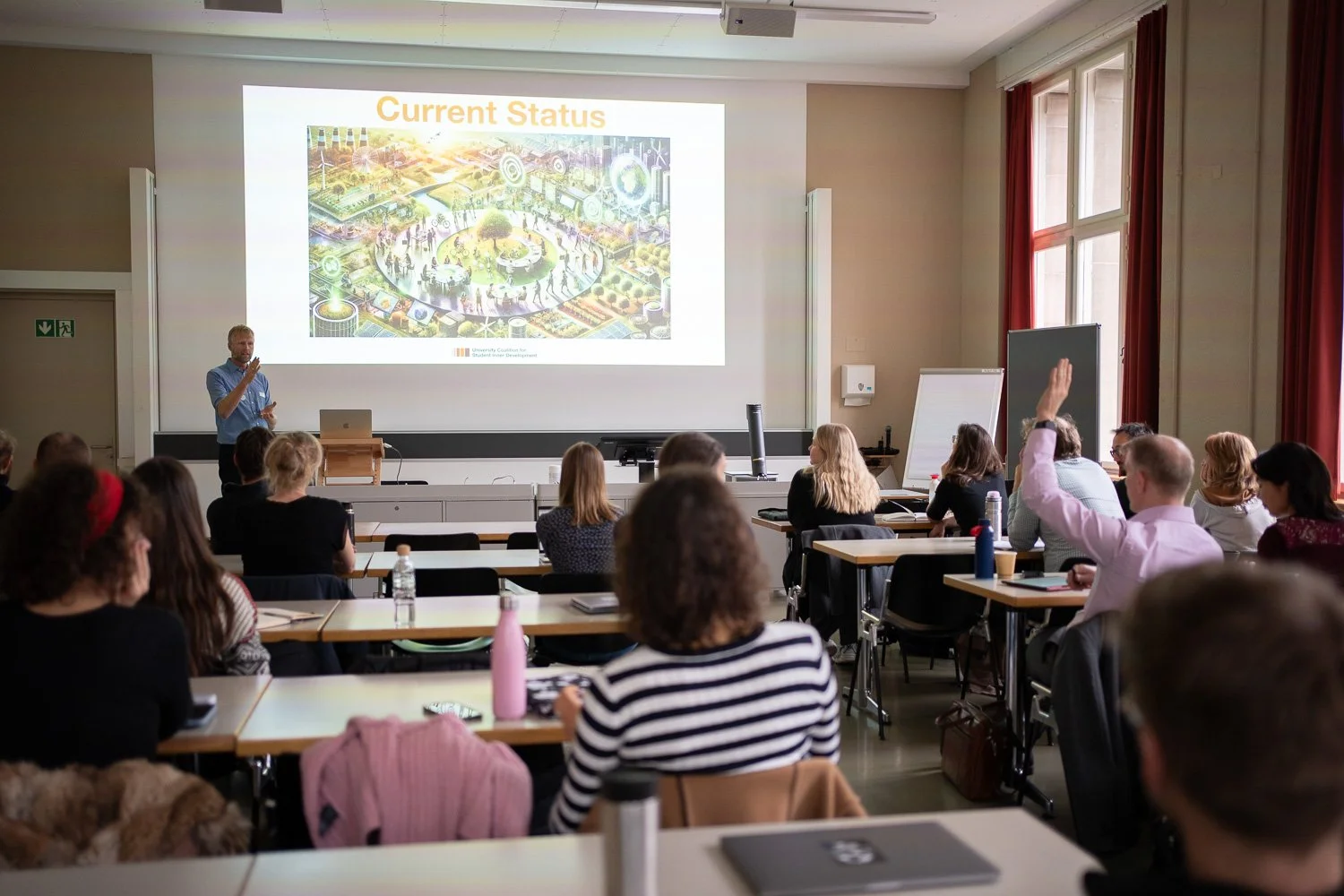 A classroom or conference room filled with people attending a presentation. The presenter is standing by a large screen displaying a slide titled 'Current Status' with colorful graphics. One attendee has their hand raised, and others are seated at ta